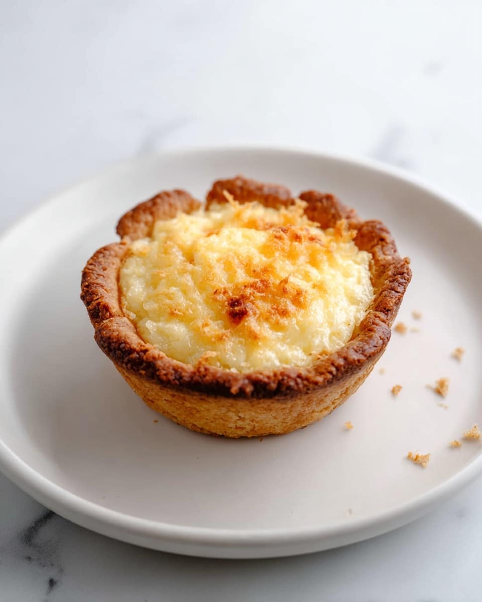 A single small tart sits in the center of a white plate. The tart has one main layer: a golden-brown crust forming a cup shape that holds a slightly raised, rough-textured filling. The filling is pale yellow with some darker golden spots on the top, suggesting it is baked. The plate rests on a white marbled surface with a few tiny crumbs scattered nearby. The photo taken with an iphone --ar 4:5 --v 7