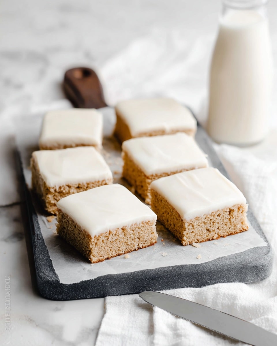 The image shows six square pieces of cake with two layers each, arranged in two rows of three on a piece of parchment paper on a dark gray cutting board. The bottom layer is a light brown, soft-looking cake with a slightly crumbly texture, while the top layer is a smooth, creamy white frosting that covers each cake square evenly. The cutting board is placed on a white cloth on a white marbled surface. In the background, on the right, there is a clear glass bottle filled with milk, and near the bottom right corner, a silver knife with some frosting on it rests on the surface. photo taken with an iphone --ar 4:5 --v 7