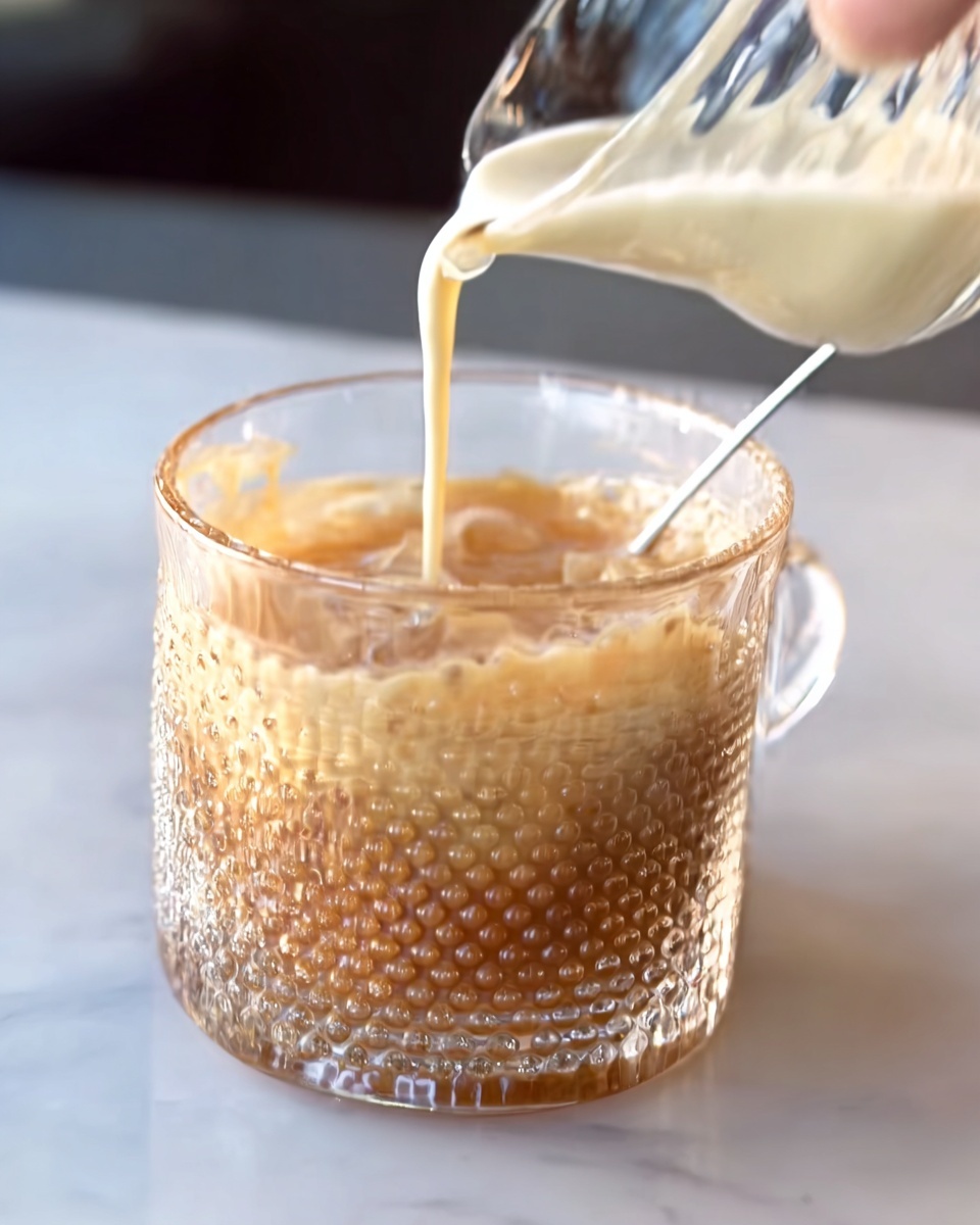A clear glass cup with a textured dotted pattern is filled with a light brown drink. A lighter cream-colored liquid is being poured from a transparent pitcher into the cup, forming a smooth top layer over the drink. The cup sits on a white marbled surface with soft lighting highlighting the texture of the drink and the cup. The scene shows only part of a woman's hand holding the pitcher. photo taken with an iphone --ar 4:5 --v 7