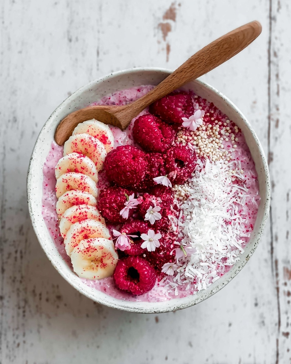 A white bowl filled with a thick pink chia pudding layer topped with a dusting of shredded white coconut spread around the edges. On one side, a row of banana slices is placed neatly, each slice sprinkled with bright red powder. Fresh red raspberries are clustered in the center, along with small light pink flower petals scattered on top. Tiny white puffed grains are scattered amidst the toppings. A wooden spoon rests inside the bowl on the upper edge. The bowl is set on a white marbled surface. Photo taken with an iphone --ar 4:5 --v 7