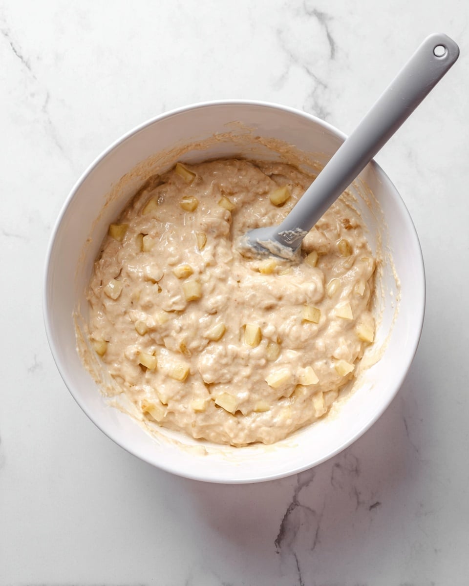 A white bowl filled with a thick, creamy light beige batter mixed with small, pale yellow apple pieces scattered evenly throughout. A metal spatula with a grey silicone head rests inside the bowl, partially covered in the batter. The bowl sits on a white marbled surface, adding a clean and bright background to the image. Photo taken with an iphone --ar 4:5 --v 7