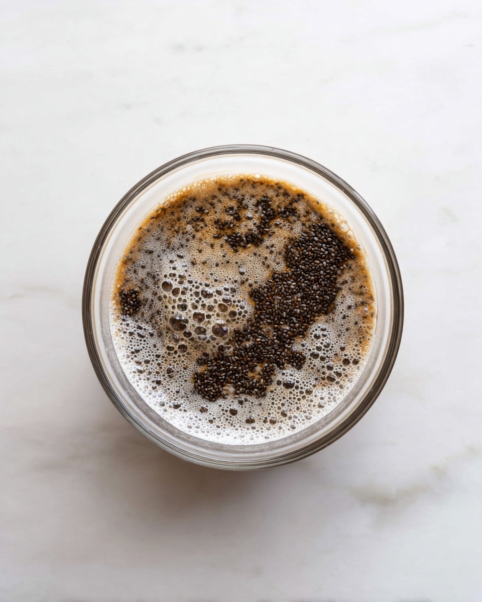 A clear glass bowl sits on a white marbled surface, filled with a mixture that has two main layers: at the bottom, a light brown liquid with some foam bubbles on top, and scattered unevenly across the surface are many small, dark brown to black chia seeds, some clustered together creating textured patches against the lighter liquid. The bowl’s smooth transparent texture shows the contents clearly from above. photo taken with an iphone --ar 4:5 --v 7