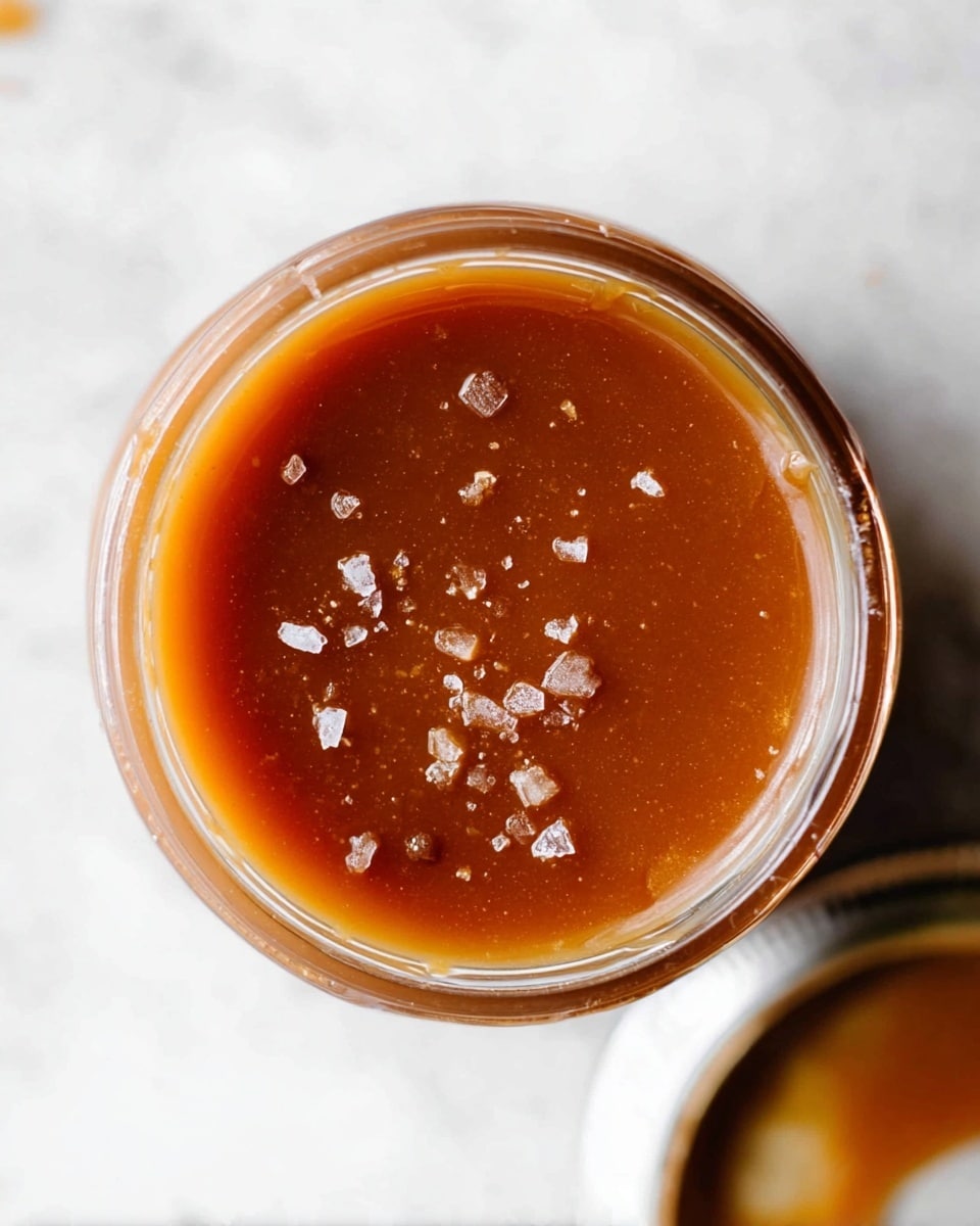 The image shows a close-up top view of a glass jar filled with smooth caramel sauce, which is deep amber-brown in color. The surface of the caramel has small chunks of coarse sea salt crystals sprinkled unevenly, adding texture and shine. The jar is sitting on a white marbled surface, and part of its lid is visible on the side. The caramel inside looks thick and glossy, with no visible layers or separation. photo taken with an iphone --ar 4:5 --v 7