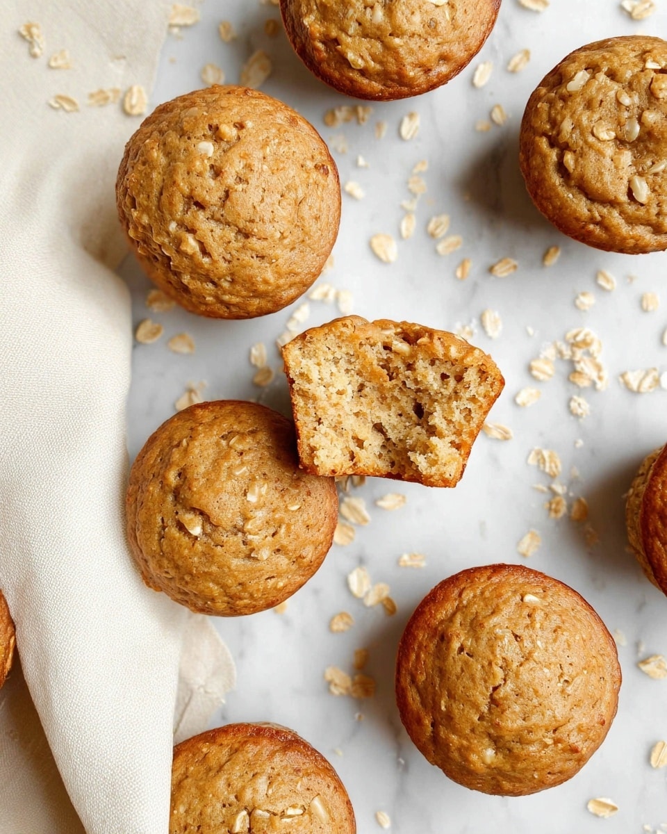 The image shows seven round oatmeal muffins with a light golden brown color and a slightly cracked top texture. One muffin is in the center with a bite taken out, showing a soft, moist inside with visible oats mixed in. The muffins are placed on a white marbled surface scattered lightly with oat flakes around them. A cream-colored cloth is partially visible on the left side. The overall arrangement looks casual and inviting. photo taken with an iphone --ar 4:5 --v 7