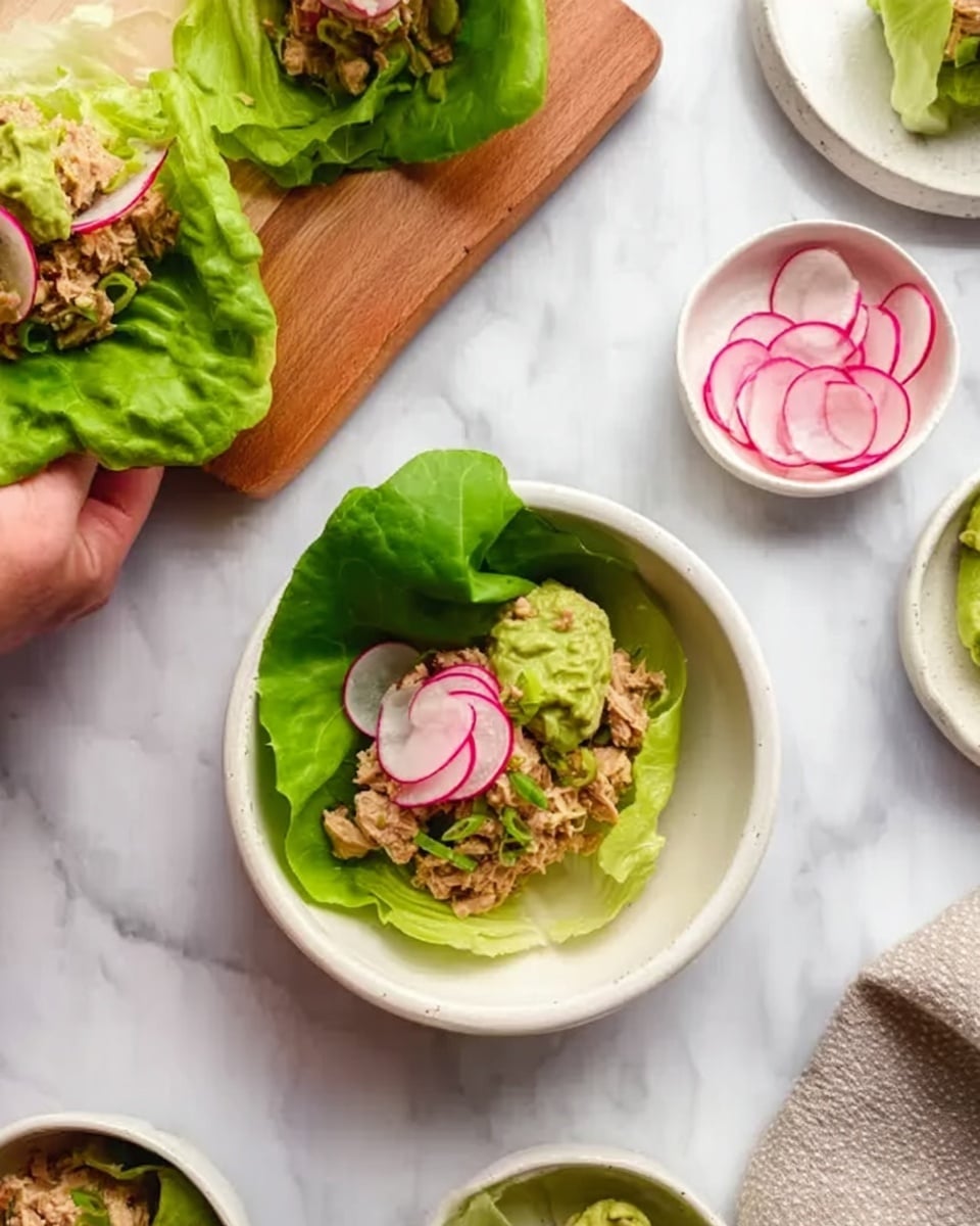 The image shows a white bowl with a single large green lettuce leaf inside. On the lettuce leaf, there is a layer of light brown chunky tuna salad mixed with small pieces, topped with light green creamy guacamole and thinly sliced pink pickled onions. Thinly sliced round radishes are scattered on top as garnish. The bowl is placed on a white marbled surface, surrounded by more white bowls with lettuce leaves and tuna salad, a small white plate with thin radish slices, and a white bowl with extra tuna salad. A woman's hand is holding a wooden serving board with another lettuce wrap in the upper left corner. Photo taken with an iphone --ar 4:5 --v 7