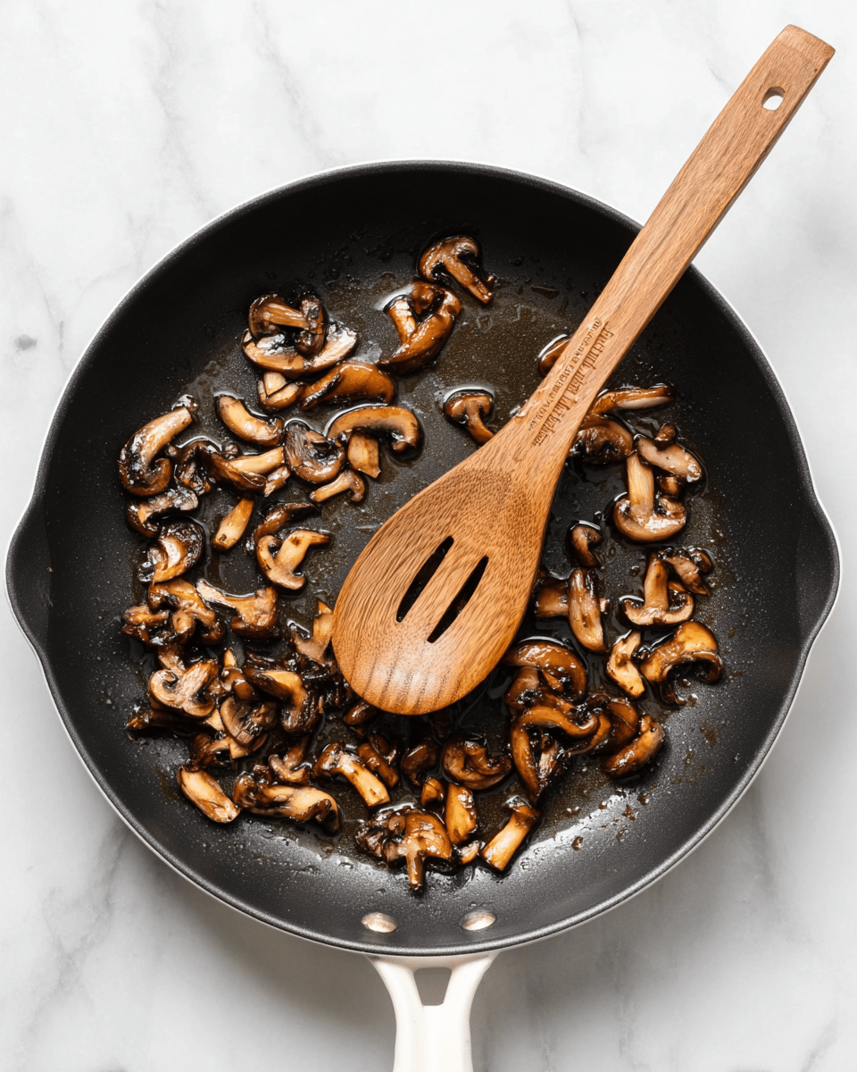 A black pan with a white handle sits on a white marbled surface, filled with small browned mushroom pieces scattered evenly across the pan's surface. The mushrooms have a shiny, slightly wet texture with golden and dark brown tones showing their cooked state. A wooden spoon with a notched tip rests diagonally inside the pan, its surface glossy with oil droplets. The scene is clean and simple, focusing on the pan and its contents. photo taken with an iphone --ar 4:5 --v 7