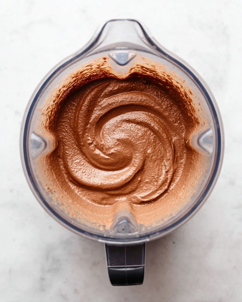 The image shows a blender with a thick, smooth brown mixture inside, which has a creamy texture and a slight swirl pattern on the surface. The blender container is transparent, revealing the chocolatey, rich mixture evenly spread inside. The blender is set on a white marbled surface, providing a clean and simple background. photo taken with an iphone --ar 4:5 --v 7