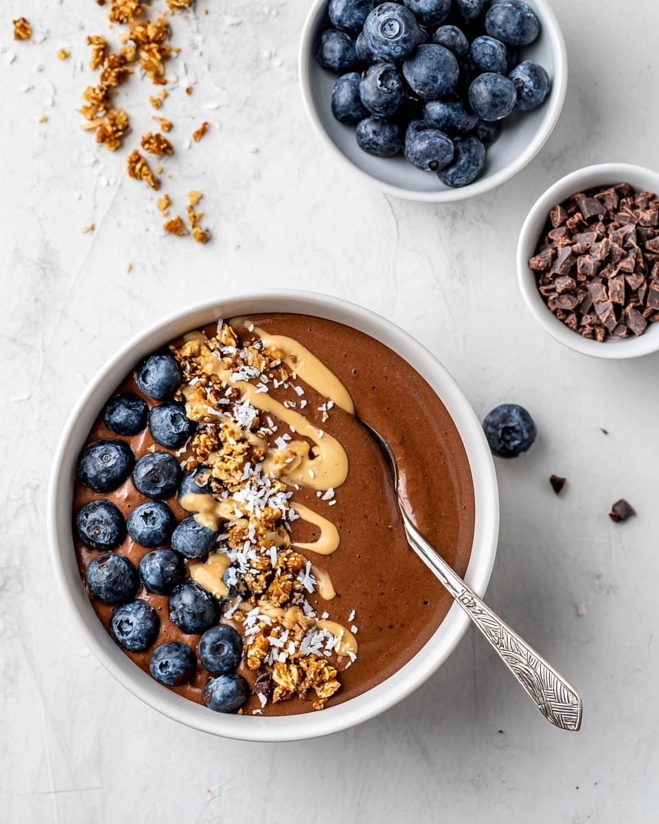 The image shows a white bowl filled with a smooth, rich brown chocolate smoothie or pudding. On top, a layer of dark blue blueberries is scattered along one side, mixed with clusters of golden brown granola and sprinkled with small bits of pale coconut flakes and seeds. Light creamy tan drizzle runs over the toppings, adding contrast. A patterned silver spoon rests inside the bowl, leaning on the edge. Nearby are two small white bowls, one with bright fresh blueberries and the other with small pieces of brown cacao nibs, all against a white marbled surface. Photo taken with an iphone --ar 4:5 --v 7