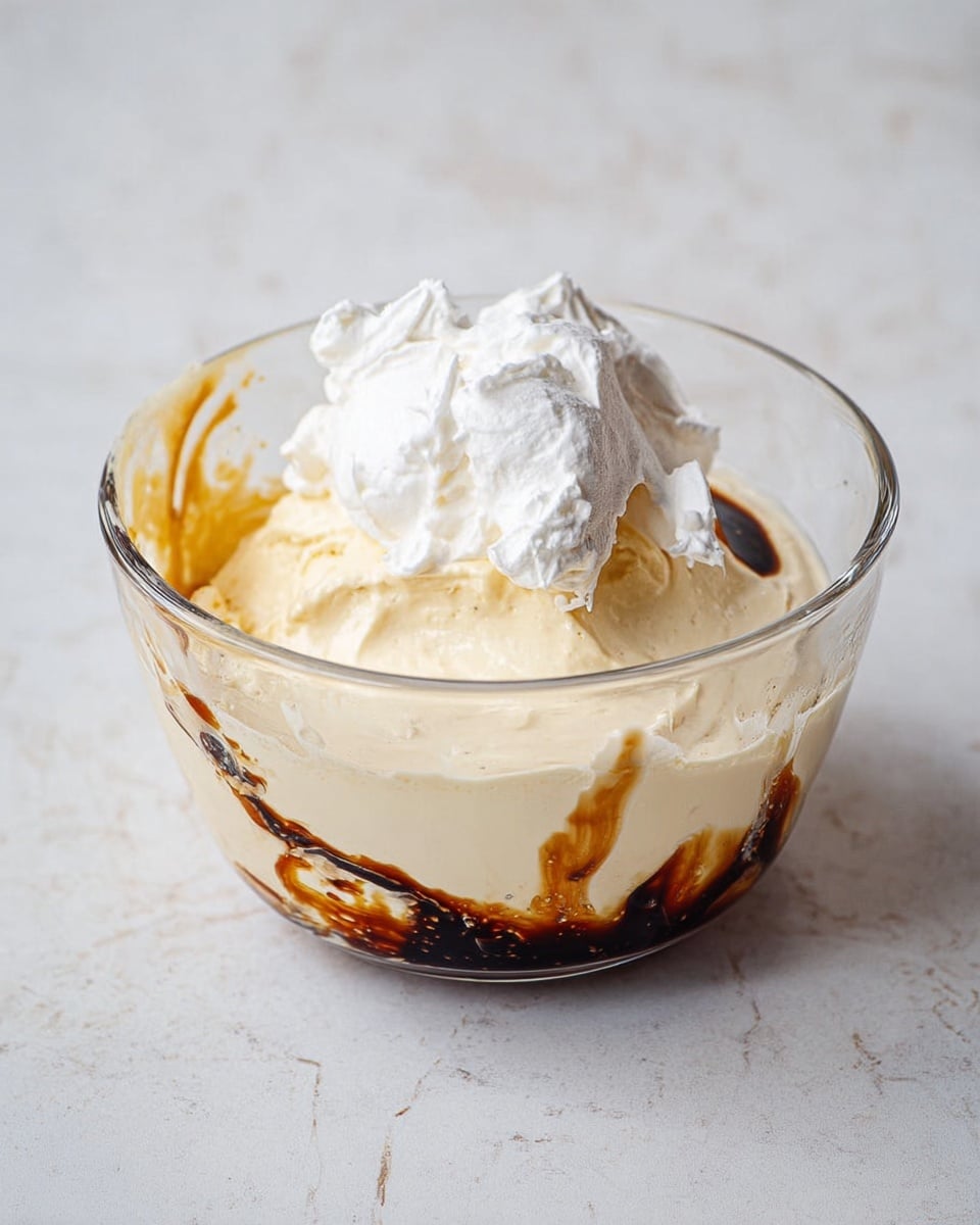 A clear glass bowl sits on a white marbled surface, containing three main layers: a large creamy pale yellow layer that looks smooth and thick, a smaller dollop of white whipped cream on top, and a dark brown syrup pooling at the edge with some uneven black streaks mixed in. The textures vary between the smooth cream, fluffy whipped cream, and the glossy syrup. Photo taken with an iphone --ar 4:5 --v 7