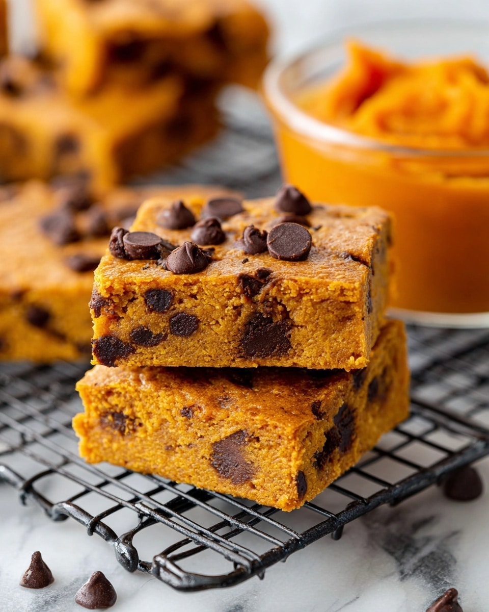 The image shows two square pumpkin bars stacked on top of each other on a black metal cooling rack over a white marbled surface. The bars have an orange-brown color with a soft, moist texture, and are filled with dark chocolate chips scattered inside and on top. The top bar is tilted slightly to show its thickness and moist inside with chocolate chips embedded. In the background, more pumpkin bars are visible on the rack, and a clear glass bowl of smooth, bright orange pumpkin puree sits slightly blurred on the white marbled surface. photo taken with an iphone --ar 4:5 --v 7