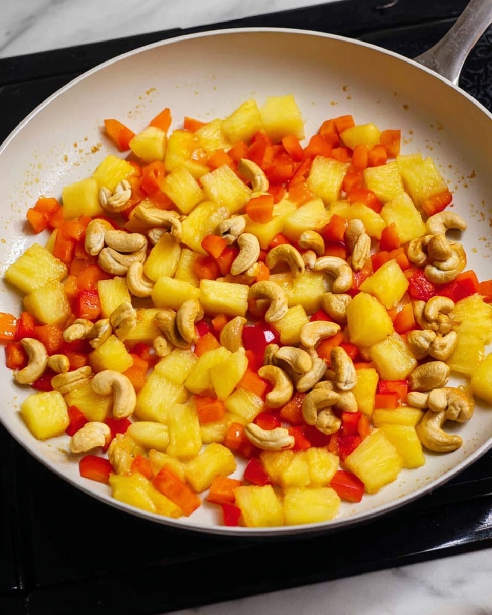 A close-up view of a white frying pan filled with three main ingredients evenly spread in one layer: small yellow pineapple chunks with a juicy texture, bright orange-red bell pepper pieces with a smooth surface, and light tan cashew nuts with a curved shape and a slightly rough texture. The pan sits on a black stovetop, and the background features a white marbled surface. The colors are vibrant and fresh, highlighting the dry cooking process. Photo taken with an iphone --ar 4:5 --v 7