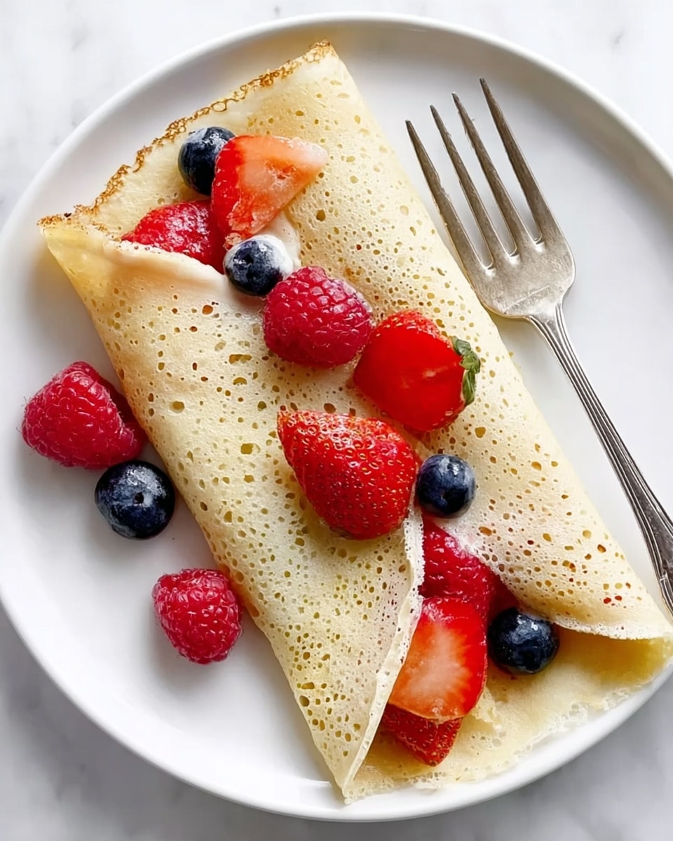 A white plate holds one thin folded crepe with a pale golden color and a light, slightly bubbly texture. The crepe is rolled around a mix of fresh berries, including bright red raspberries, halves of vibrant red strawberries with green tops, and round, dark blue blueberries. The berries peek out from the open ends of the crepe. A silver fork rests on the plate near the crepe. The whole scene is set on a white marbled surface. photo taken with an iphone --ar 4:5 --v 7