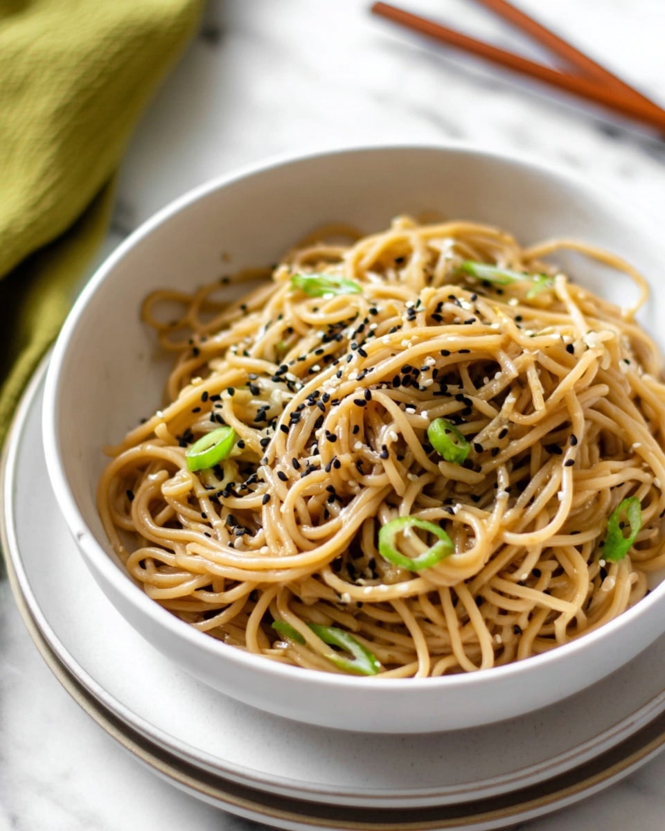 The image shows a white bowl filled with light brown noodles tossed in a glossy sauce. The noodles are loosely piled in the bowl, with some strands curled on top. Scattered on the noodles are small black and white sesame seeds and thin slices of bright green spring onions. The bowl sits on a white plate on a white marbled surface, with a blurred green cloth and wooden chopsticks in the background. photo taken with an iphone --ar 4:5 --v 7