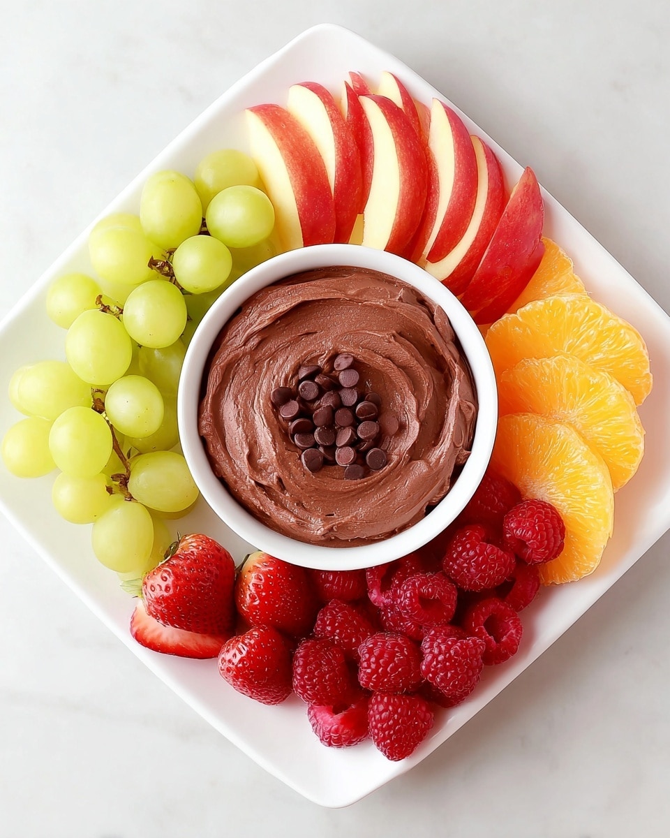 A white square plate on a white marbled surface holds a round white bowl in the center filled with smooth, swirled chocolate dip topped with small chocolate chips scattered on top. Around the bowl, there are five different fruit sections arranged in neat clusters: bright green grapes on the top left, red strawberries cut in half on the top right, thin red and yellow apple slices on the right, orange mandarin segments on the bottom right, and red raspberries on the bottom left. The colors are vibrant and fresh, creating a balanced and colorful fruit and dip arrangement. photo taken with an iphone --ar 4:5 --v 7