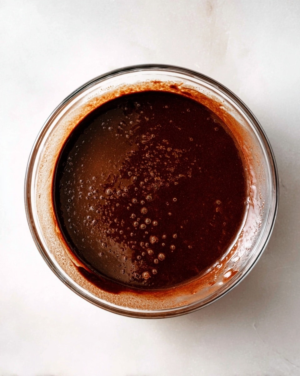 A clear round glass bowl is placed on a white marbled surface, filled with a thick, dark brown chocolate sauce that has a smooth texture with some small air bubbles and slight unevenness on the surface. The sauce layer fills about half of the bowl, with some sauce dripped on the sides just above the surface level. Photo taken with an iphone --ar 4:5 --v 7