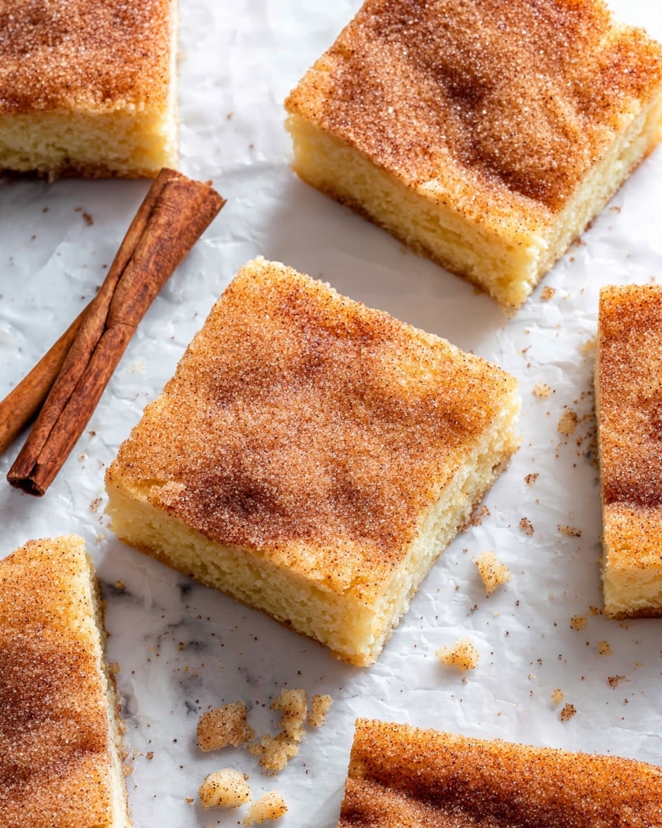 The image shows several square pieces of cinnamon sugar cake arranged on white parchment paper over a white marbled surface. Each piece has two layers: a light golden yellow base that looks soft and moist, topped with a thin crust sprinkled evenly with a mixture of cinnamon and sugar, creating a warm brown, slightly grainy texture. Near the center-left, two cinnamon sticks cross each other, adding a rustic touch. Small crumbs and sugar grains scatter lightly around the pieces, enhancing the fresh-baked look. Photo taken with an iphone --ar 4:5 --v 7