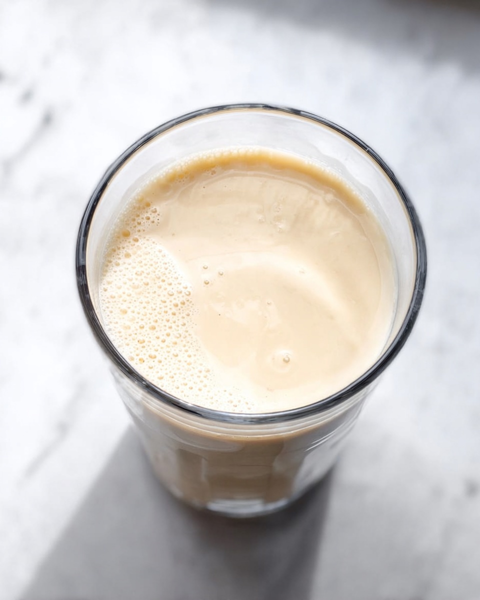 A clear cylindrical glass filled with a light beige creamy liquid that has a smooth texture with small bubbles around the edges and a few soft swirls in the middle. The glass is placed on a white marbled surface, with the top view showing the liquid's surface clearly. The background and surrounding area are bright and clean, highlighting the creamy drink inside the glass. Photo taken with an iphone --ar 4:5 --v 7