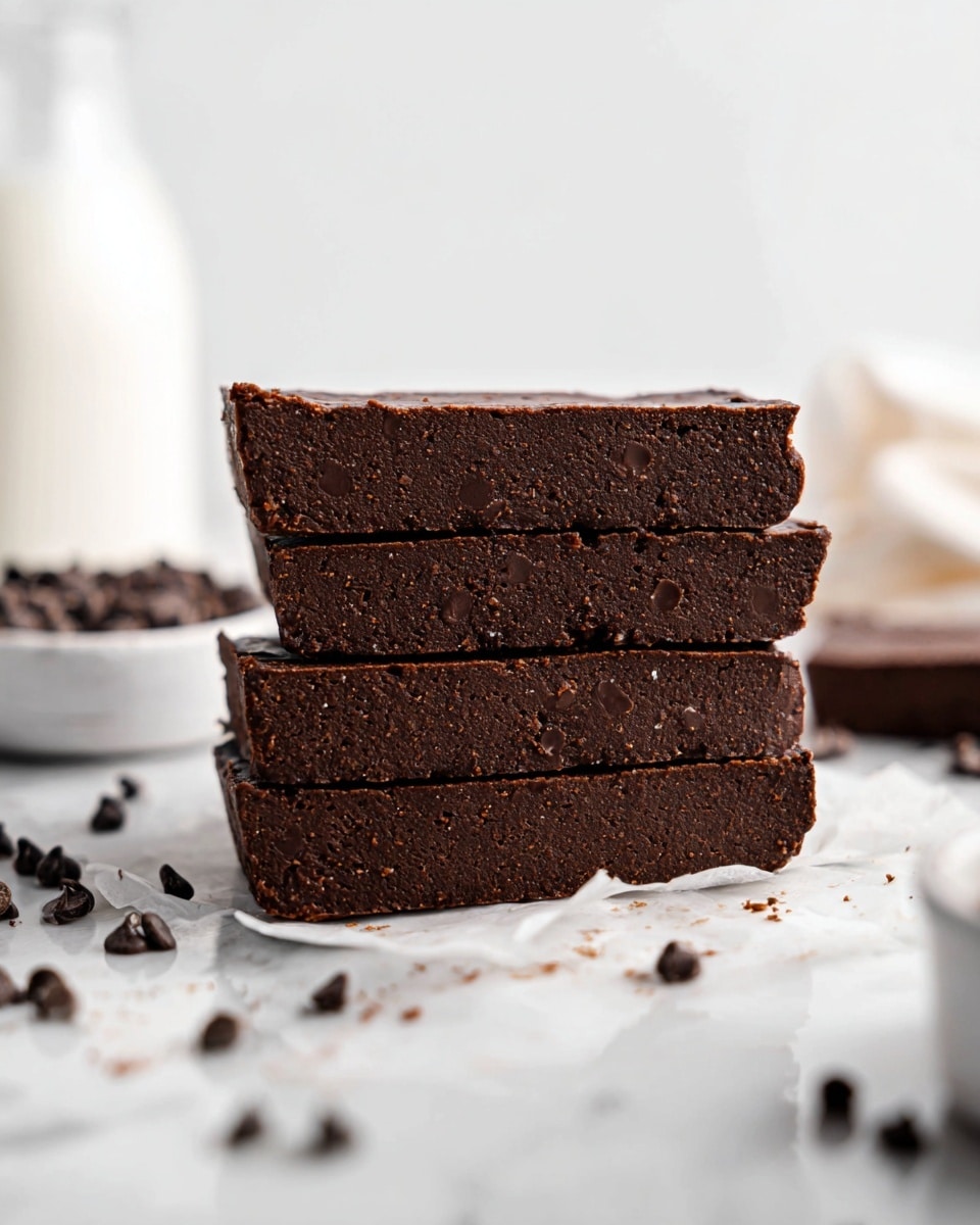 Four thick, dark brown chocolate bars are stacked on top of each other, showing a dense and slightly crumbly texture with small chocolate chips embedded in them. The bars are placed on a white marbled surface with scattered chocolate chips around, adding texture and depth. In the background, blurry white bowls and a milk bottle add a soft contrast to the rich chocolate. The overall look is simple and clean, focusing on the three-dimensional layers of the chocolate bars. photo taken with an iphone --ar 4:5 --v 7