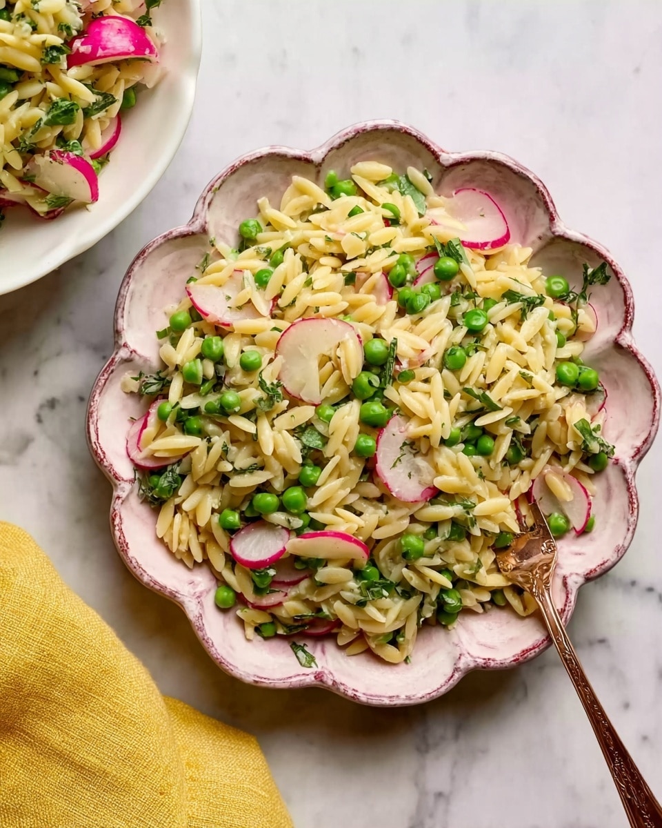 The image shows a white scalloped bowl and a white round plate with a pink rim, both filled with a colorful orzo salad. The salad has three layers mostly mixed together: pale yellow orzo pasta, bright green peas, and thin, light pink radish slices. The pasta looks soft and glossy, peas are round and fresh, and radishes are thin and slightly translucent. There is a bronze fork lying on the plate partially resting on the salad. The background is a white marbled surface, and a yellow cloth is partly visible at the bottom edge. Photo taken with an iphone --ar 4:5 --v 7