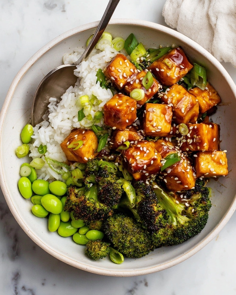A white bowl contains a layered dish with three main parts: a portion of white rice on the left with a shiny silver spoon partially scooping it, bright green edamame beans scattered mostly near the bottom layer, and roasted broccoli florets with green stems placed around the right side and top, showing some charred texture. On top, golden-brown tofu cubes coated in a shiny brown sauce sprinkled with white sesame seeds form the central layer, along with pieces of green onion mixed among the tofu. The bowl sits on a white marbled surface. photo taken with an iphone --ar 4:5 --v 7