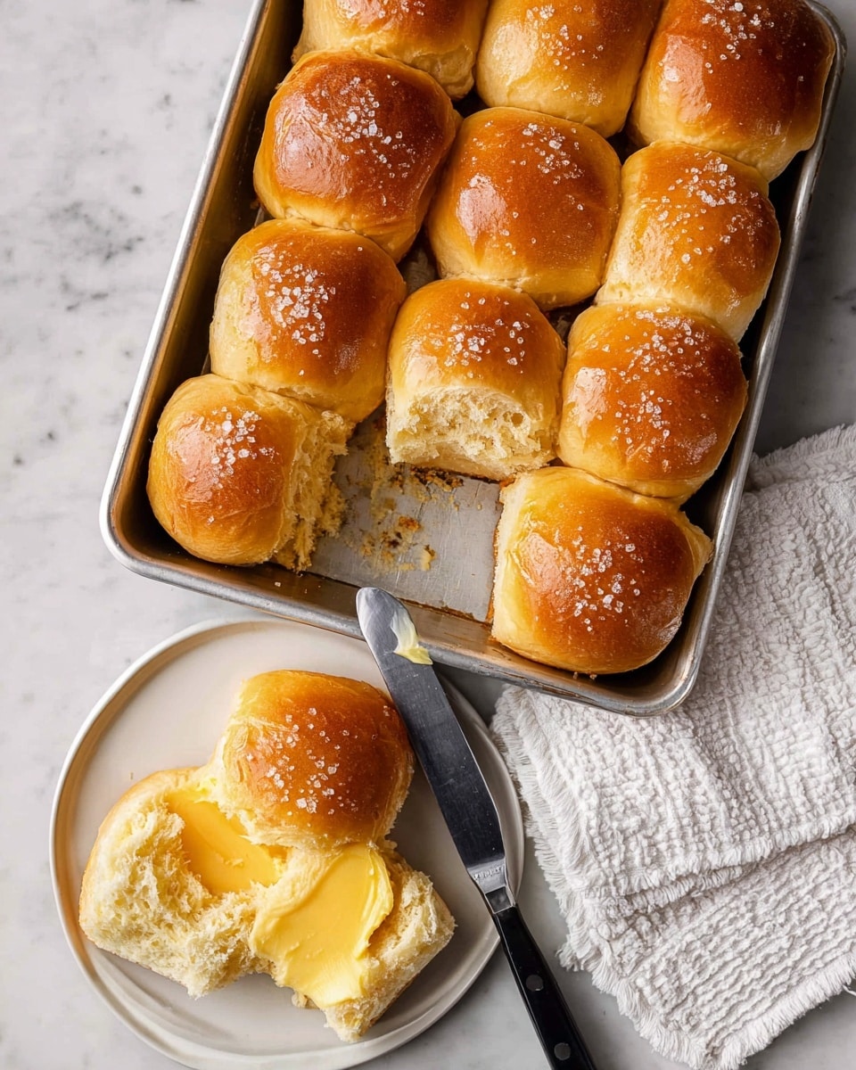 A tray filled with 11 golden brown dinner rolls, soft and shiny on top with a slight glaze and sprinkled lightly with coarse salt. One roll is torn open to show a fluffy, light inside. Below the tray, a white plate holds one of the rolls, split open and spread with creamy yellow butter. A butter knife with a black and white handle rests on the plate’s edge. The tray and plate sit on a white marbled surface, and a white textured cloth is placed near the tray photo taken with an iphone --ar 4:5 --v 7