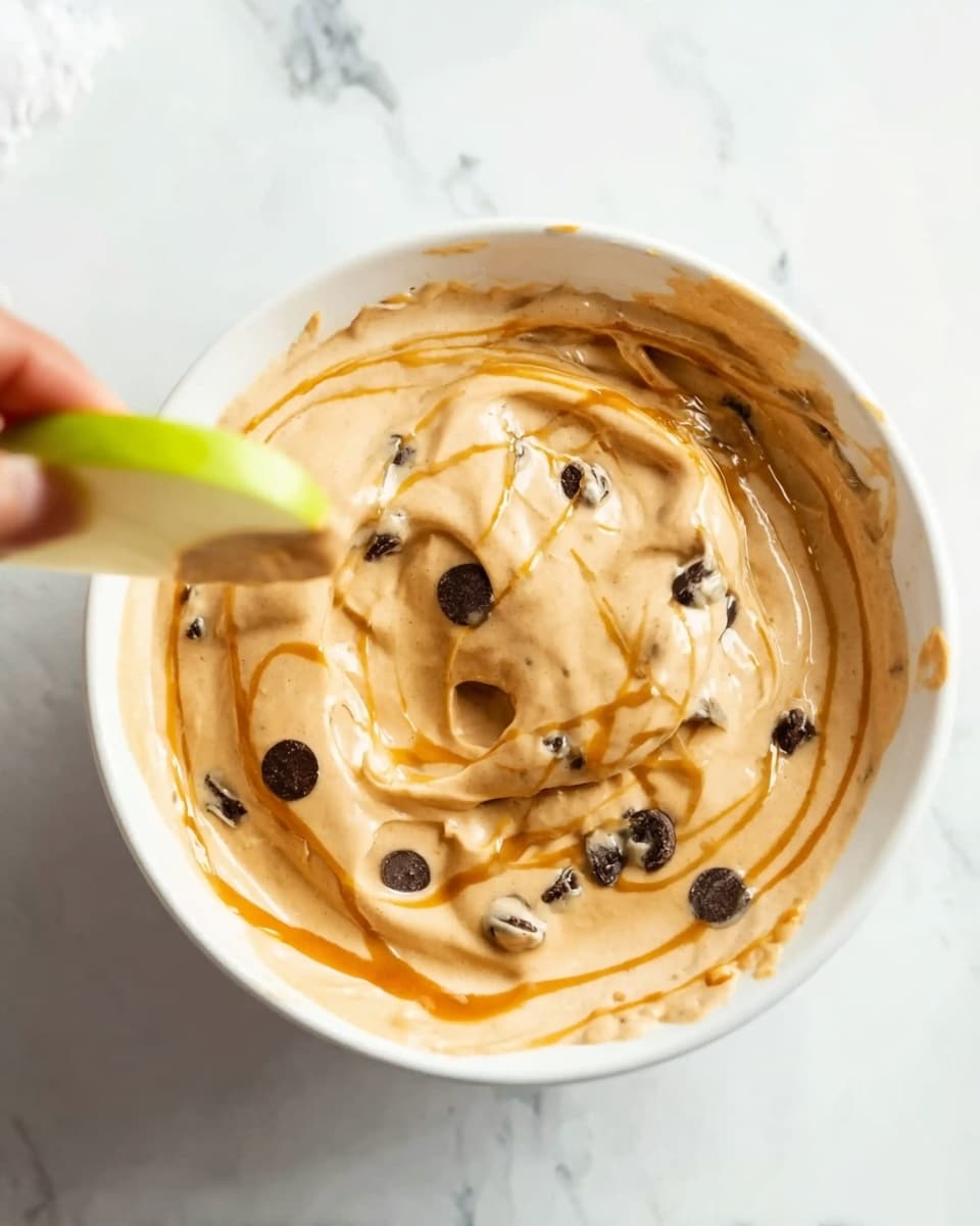 A close-up view of a white bowl filled with a creamy, light brown mixture that has a smooth texture with some dark chocolate chips scattered throughout. The creamy layer is swirled with thin, golden caramel streaks on top. A woman's hand holds a green apple slice dipped into the mixture, lifting it above the bowl. The background and surface are white marble, keeping the focus on the bowl and the apple slice photo taken with an iphone --ar 4:5 --v 7
