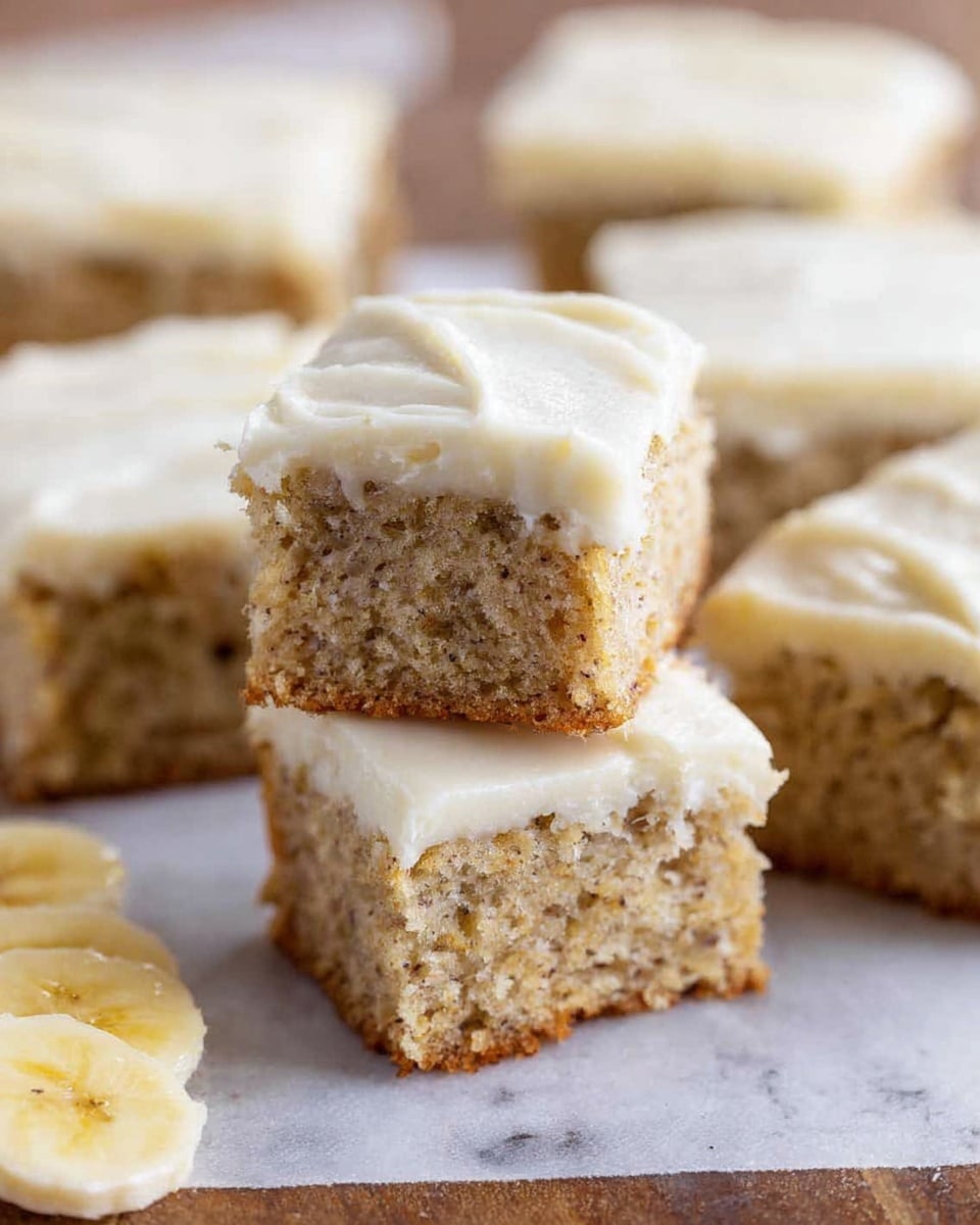 The image shows a group of small square banana cake pieces with a light brown moist texture and tiny dark specks inside, topped with a thick layer of creamy white frosting that looks smooth and slightly swirled. Three pieces are stacked in the center, one on top of two others, and more pieces are blurred in the background. The pieces rest on a white marbled surface with some sliced bananas visible on the lower left corner. Photo taken with an iphone --ar 4:5 --v 7