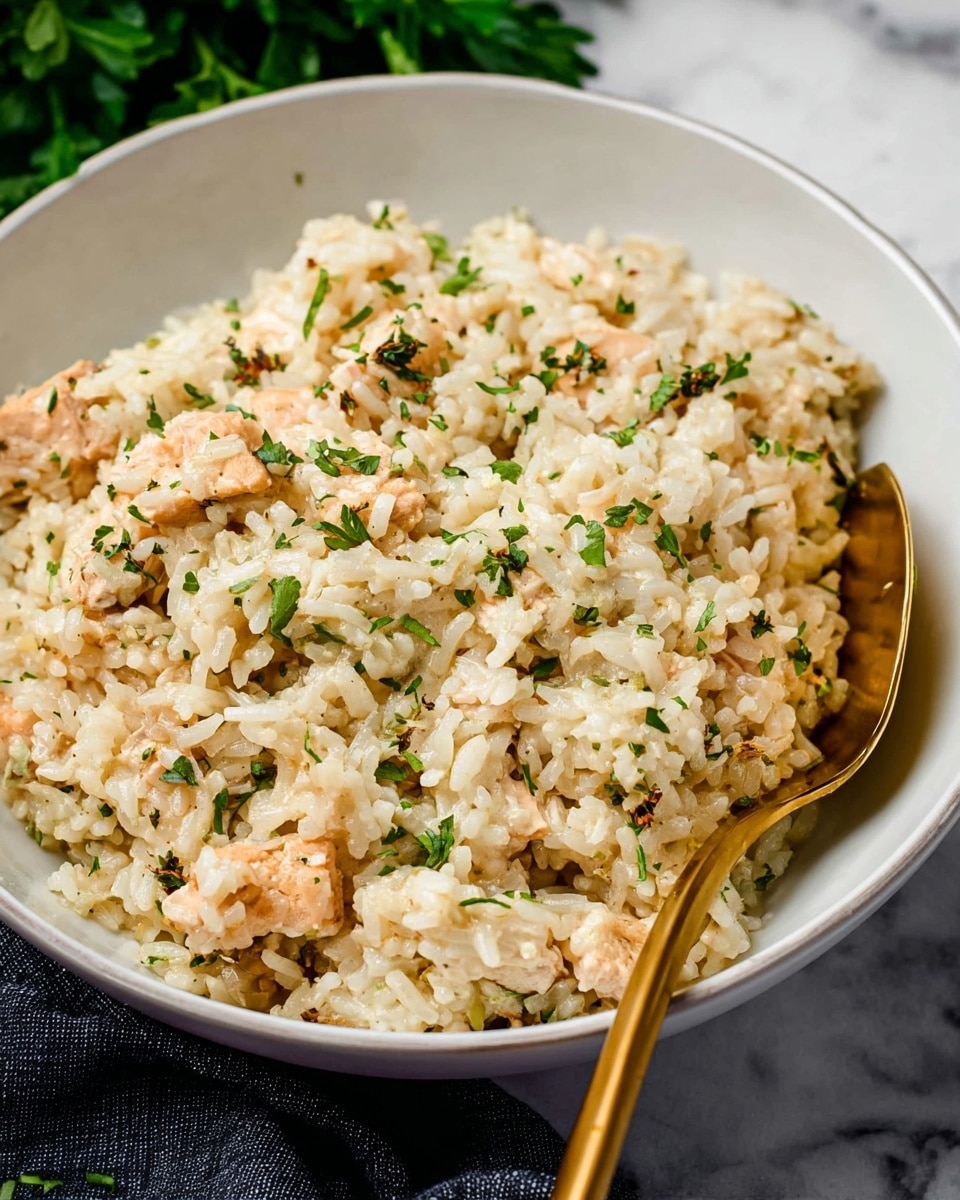 A close-up of a white deep bowl filled with creamy rice mixed with small pieces of light brown cooked chicken and finely chopped green herbs sprinkled on top. The rice looks soft and coated evenly with a light cream sauce, and a gold spoon is partially buried in the rice on the right side of the bowl. The bowl sits on a white marbled surface with a dark cloth and green leaves blurred in the background. Photo taken with an iphone --ar 4:5 --v 7