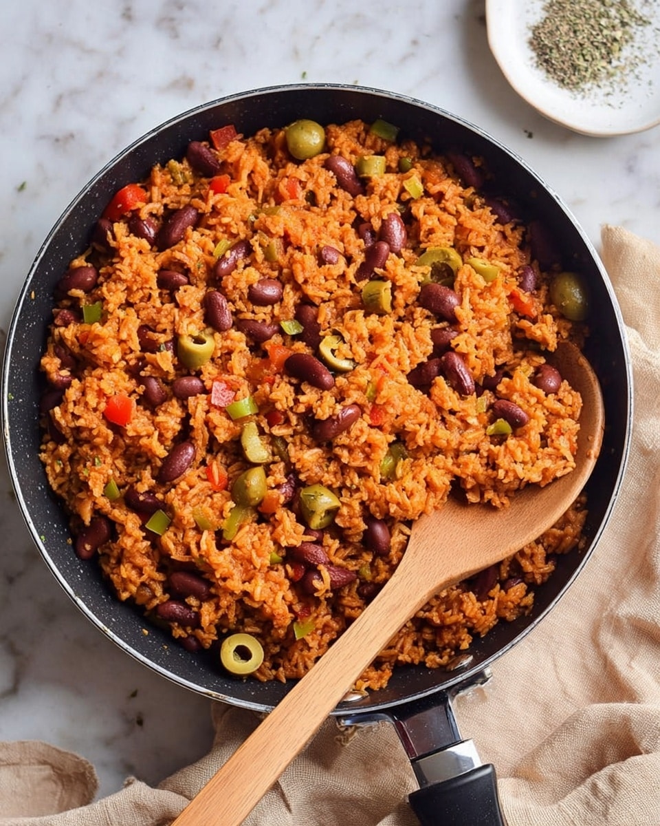 A black pan filled with cooked rice mixed with red beans, small green olive slices, and diced red and green vegetables. The rice is orange in color, showing it is well seasoned, with the beans and vegetables spread evenly throughout. A wooden spoon is placed inside the pan with its handle extending outwards. The pan sits on a white marbled surface with a beige cloth nearby and a white plate with seasoning in the top right corner. Photo taken with an iphone --ar 4:5 --v 7