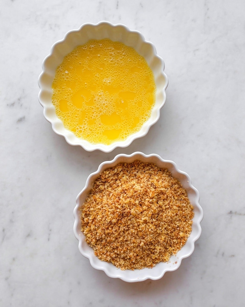 Two white scalloped bowls are placed on a white marbled surface. The bowl on the left contains a bright yellow beaten egg mixture with bubbles on the surface. The bowl on the right is filled with a coarse, crumbly mixture of golden brown breadcrumbs with some variation in color. Both bowls are viewed from above in natural light. photo taken with an iphone --ar 4:5 --v 7