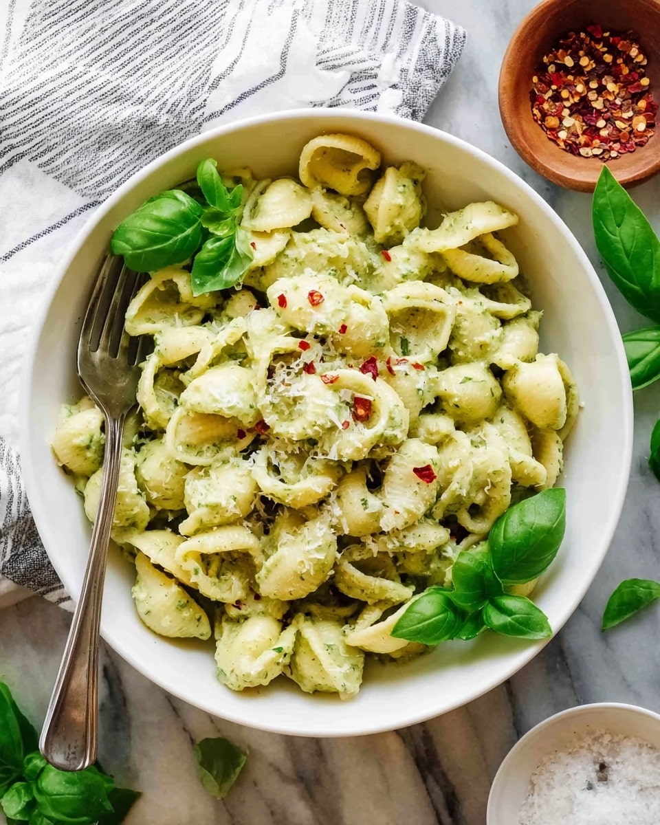 A white bowl filled with small shell-shaped pasta tossed in a creamy light green sauce, topped with some finely grated cheese and a few red chili flakes scattered on top. Fresh green basil leaves are placed around the edges and in the center of the pasta. A silver fork is resting inside the bowl on the left side. The bowl is on a white marbled surface with fresh basil leaves nearby and a small white cup of pepper flakes and salt to the sides. A striped white cloth napkin is partly visible at the top left corner. Photo taken with an iphone --ar 4:5 --v 7