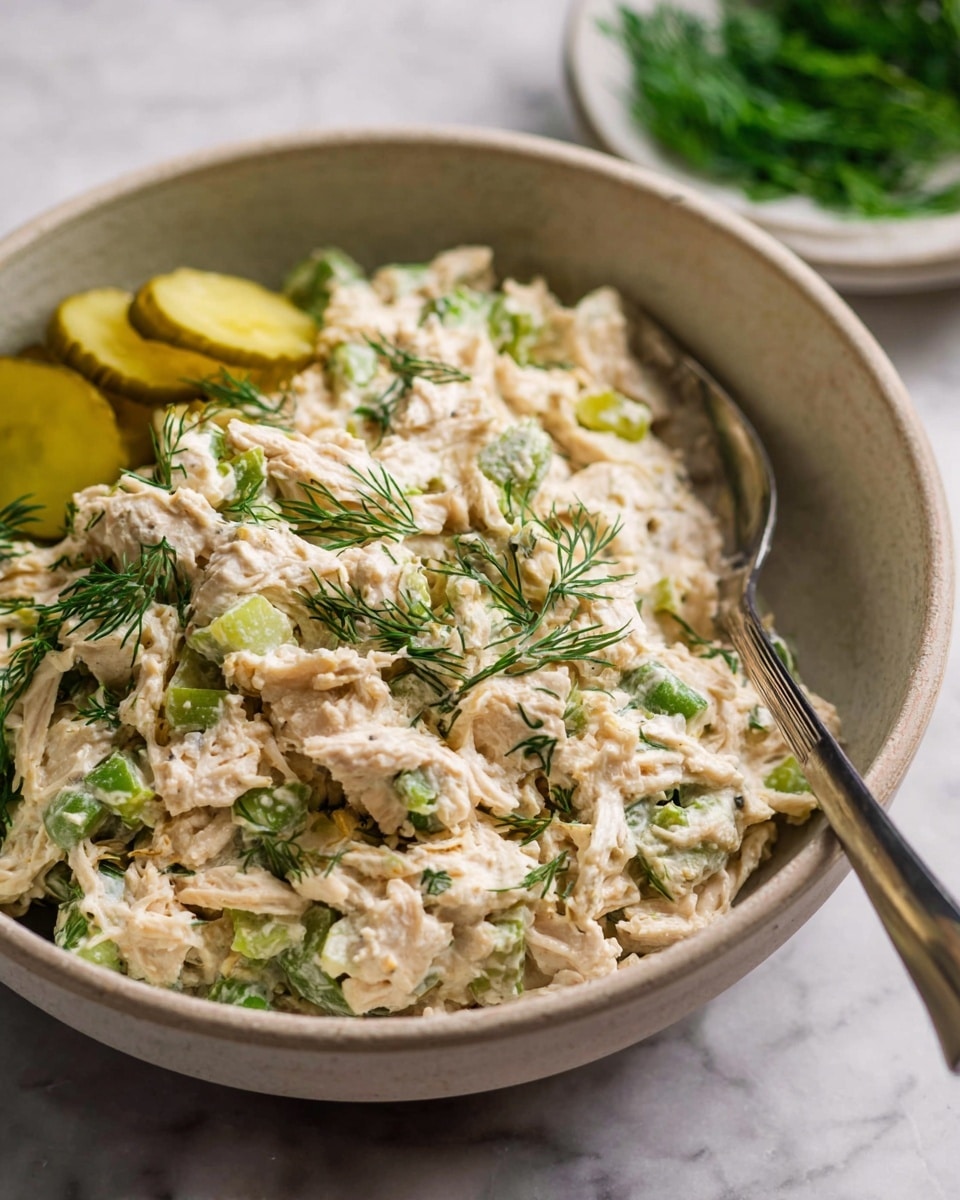 A beige bowl filled with creamy chicken salad that has pieces of white shredded chicken and bits of green celery mixed in. The salad is speckled with green dill herbs scattered on top and mixed inside. There are three thin slices of pickles placed on one side of the bowl. A silver spoon is resting inside the bowl, partly buried in the salad. In the blurry background, a white bowl holds more fresh green dill. The bowl sits on a white marbled surface photo taken with an iphone --ar 4:5 --v 7