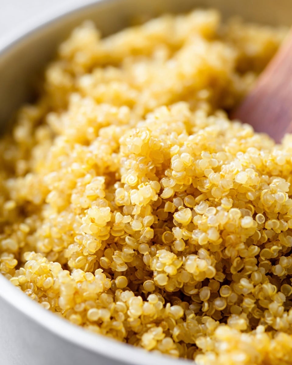 A close-up view of cooked quinoa grains, shown in a white bowl with a wooden spoon partly visible in the background. The quinoa grains are small, round, and tightly packed, with a light golden yellow color and a slightly fluffy texture. The grains appear soft and moist, with some showing their characteristic tiny spiral curls. The white marbled texture surface is blurred out in the background, focusing on the even, textured layer of quinoa filling the bowl. photo taken with an iphone --ar 4:5 --v 7