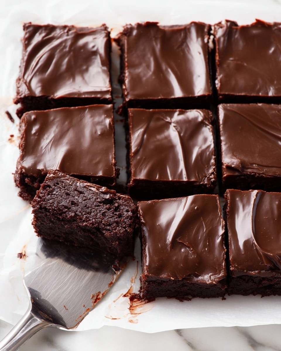 The image shows nine square pieces of chocolate dessert with a shiny, smooth dark chocolate layer on top. The top layer looks moist and slightly wrinkled, covering a dense, dark chocolate base underneath. One piece has been taken out, revealing a thick, fudgy texture with some chocolate bits around it. The dessert pieces rest on white parchment paper, and a shiny metal spatula is lifting the piece that has a bite missing. The background is a white marbled surface. photo taken with an iphone --ar 4:5 --v 7