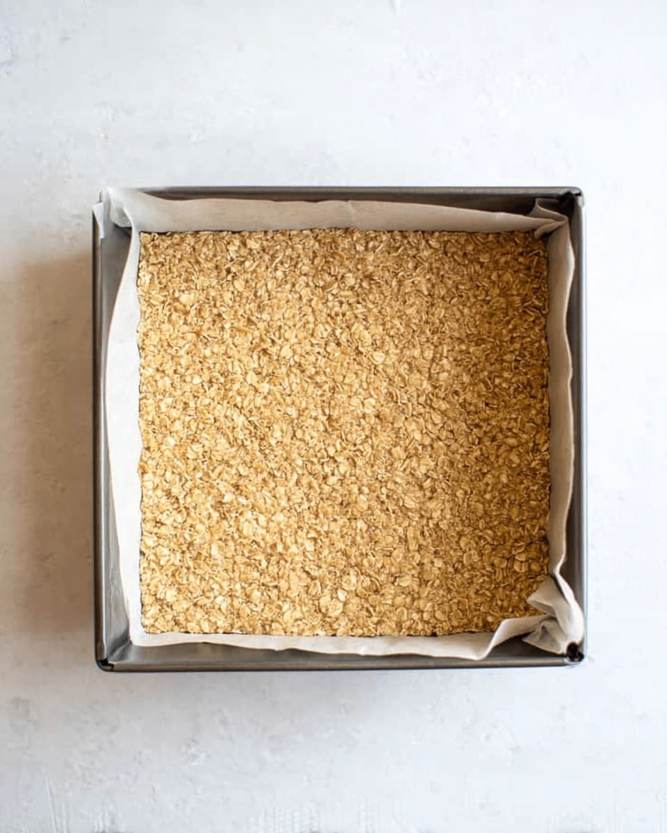 A top view shows a close-up of a square baking pan lined with white parchment paper, filled with a thick, flat layer of light brown, crumbly oat mixture that has a rough, grainy texture and uneven edges. The pan sits on a white marbled surface, emphasizing the oat layer's golden color and coarse oats spread evenly across the top. Photo taken with an iphone --ar 4:5 --v 7