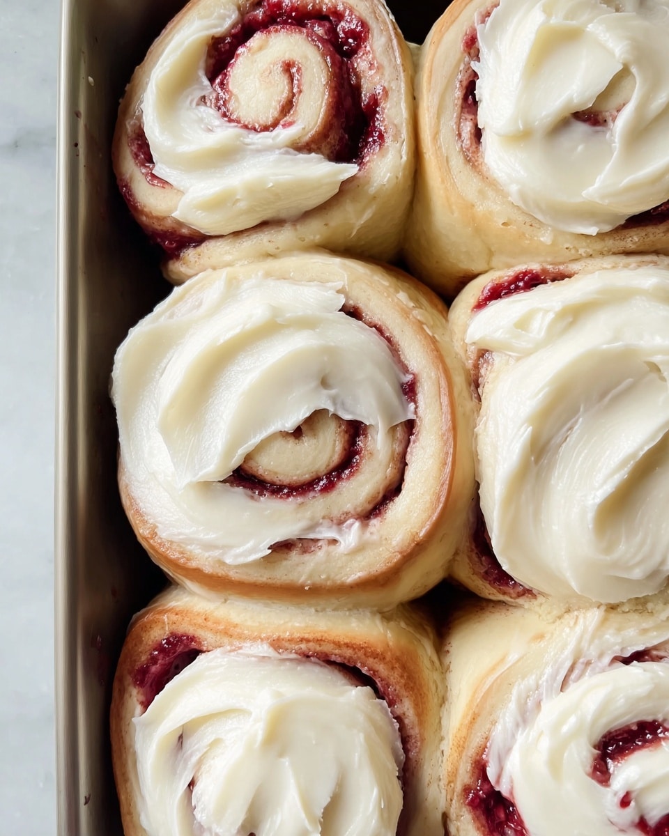 This image shows a close-up of six soft swirled buns arranged in a baking pan on a white marbled surface. Each bun is rolled with a deep red filling, visible in spirals inside the pale golden brown dough. They are topped with a thick, creamy layer of smooth white frosting, spread in wide swirls over each bun, partly covering the red filling but leaving some spiral parts exposed. The texture of the frosting appears fluffy and creamy, with gentle peaks from the spreading. The buns sit closely side by side, filling the pan evenly. Photo taken with an iphone --ar 4:5 --v 7