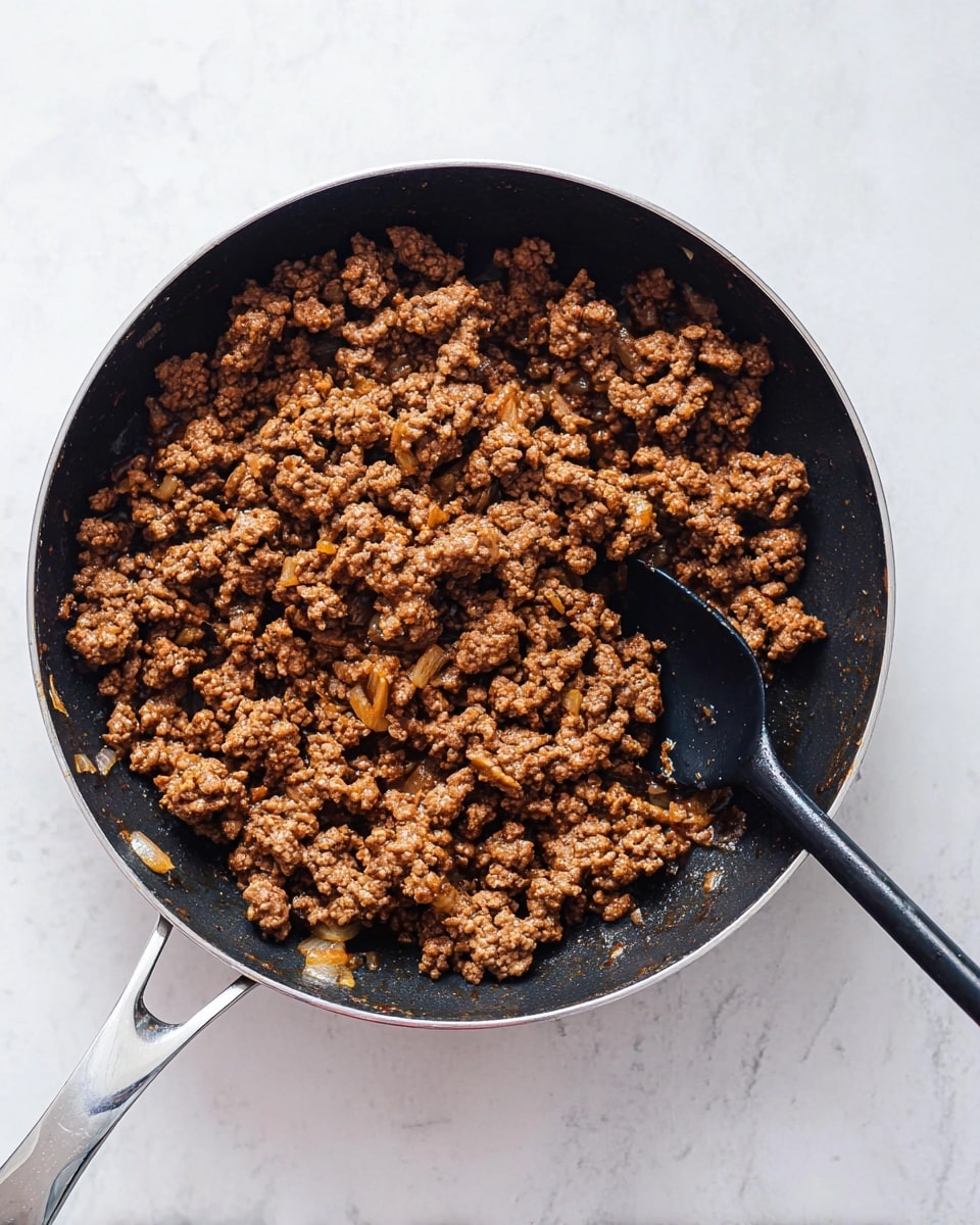 A black pan filled with cooked ground meat that is brown and crumbly with small pieces of onion mixed in, and some oil glistening on the surface. A black spoon rests inside the pan on the right side, slightly submerged in the meat. The pan handle is silver and placed on a white marbled surface. Photo taken with an iphone --ar 4:5 --v 7