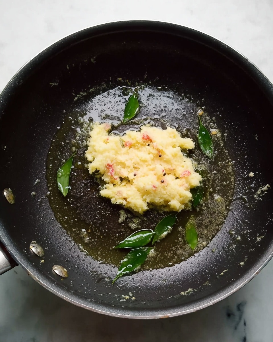 A black frying pan on a white marbled surface holds a small mound of pale yellow mashed mixture with some small bits of red and white visible inside. Around the mash, several fresh green leaves float in a slightly oily liquid, which glistens under the light. The pan's interior shows light signs of use with a smooth texture, and the whole scene has a warm, cooking-in-progress feel. photo taken with an iphone --ar 4:5 --v 7