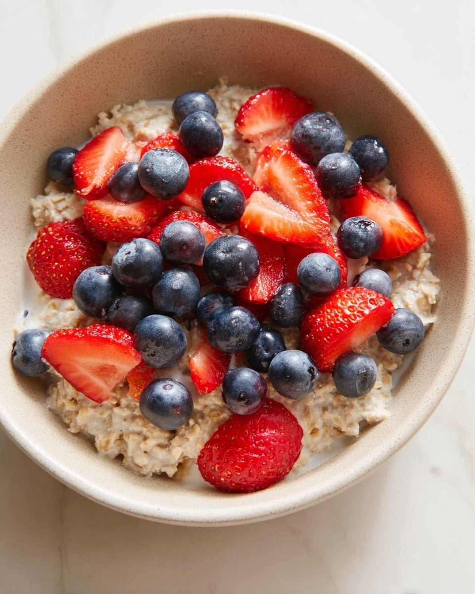 A close-up view of a beige bowl filled with creamy oatmeal as the base layer, topped with a mix of fresh blueberries and sliced strawberries. The strawberries are bright red with a juicy texture, sliced into round pieces and scattered evenly over the oatmeal. The blueberries are plump with a deep blue color, resting naturally among the strawberries. The bowl sits on a white marbled surface, showing a soft natural light that highlights the fresh colors of the fruit. Photo taken with an iphone --ar 4:5 --v 7