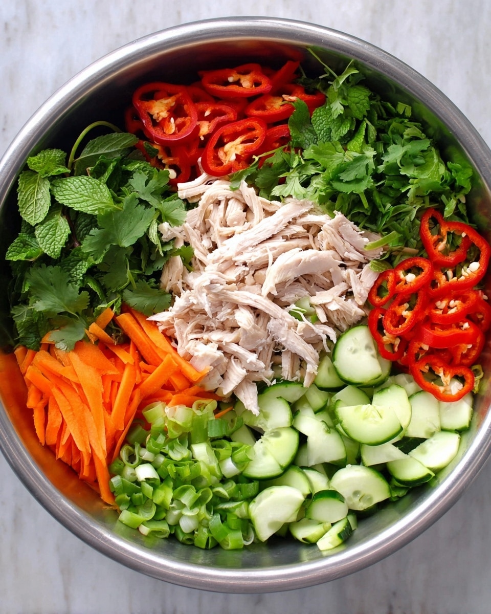 This image shows a large metal bowl filled with seven separate layers of fresh ingredients. At the center is a pile of shredded cooked chicken, light brown and white in color, surrounded by layers of green cilantro leaves at the top, thinly sliced red bell peppers to the top right, thin carrot sticks to the lower right, thin red chili rings on top of the carrots, chopped green onions at the bottom right, half-moon slices of cucumber at the bottom left, and green mint leaves to the left side. Each layer is neatly arranged side by side, showing a mix of bright green, orange, red, and white colors. The bowl is placed on a white marbled surface. photo taken with an iphone --ar 4:5 --v 7