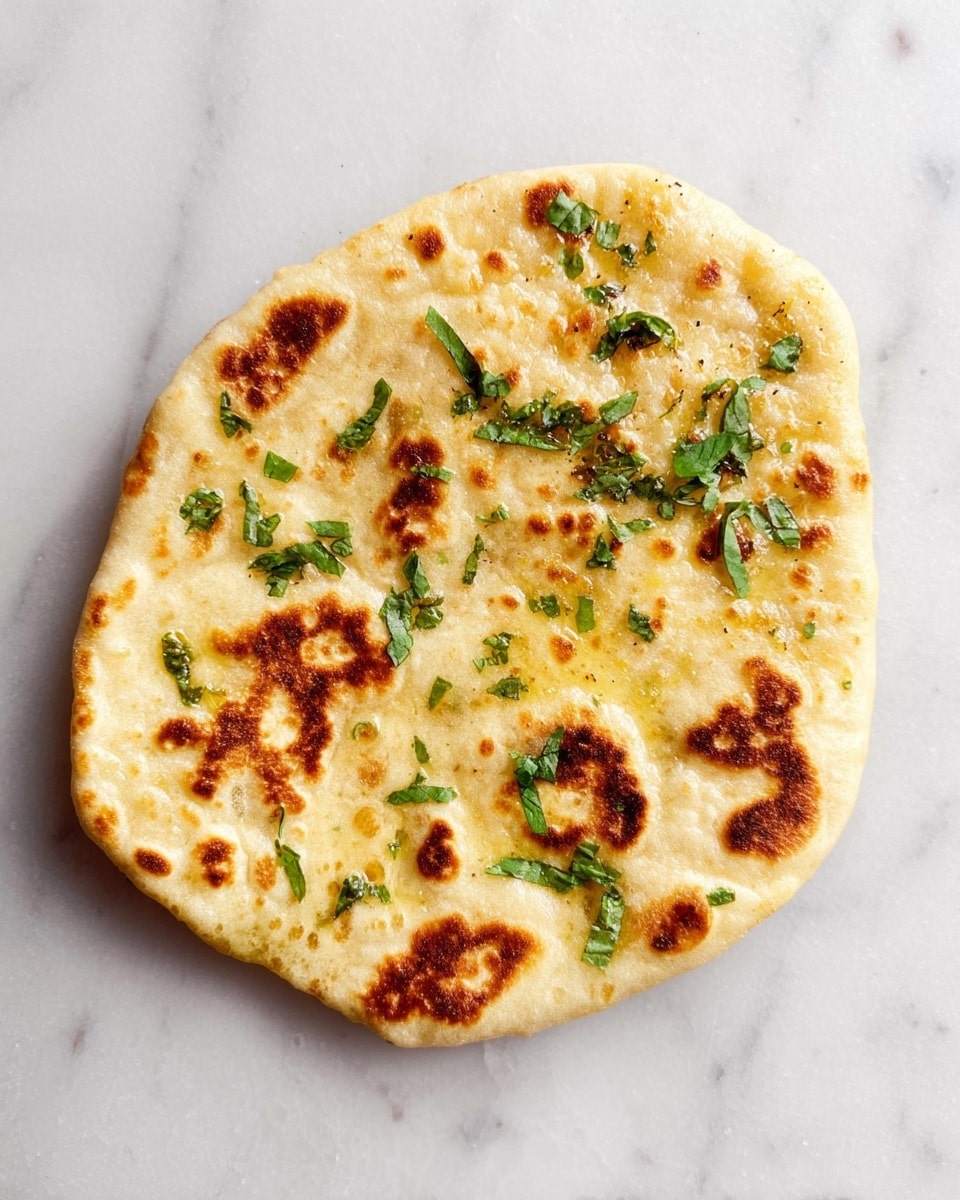 A single flat round bread with a soft, golden surface and brown toasted spots is shown directly on a white marbled surface. The bread is topped with scattered small green leaves, adding a fresh look. The texture is slightly uneven, with some bubbles and a lightly shiny finish from a brush of oil or butter. photo taken with an iphone --ar 4:5 --v 7