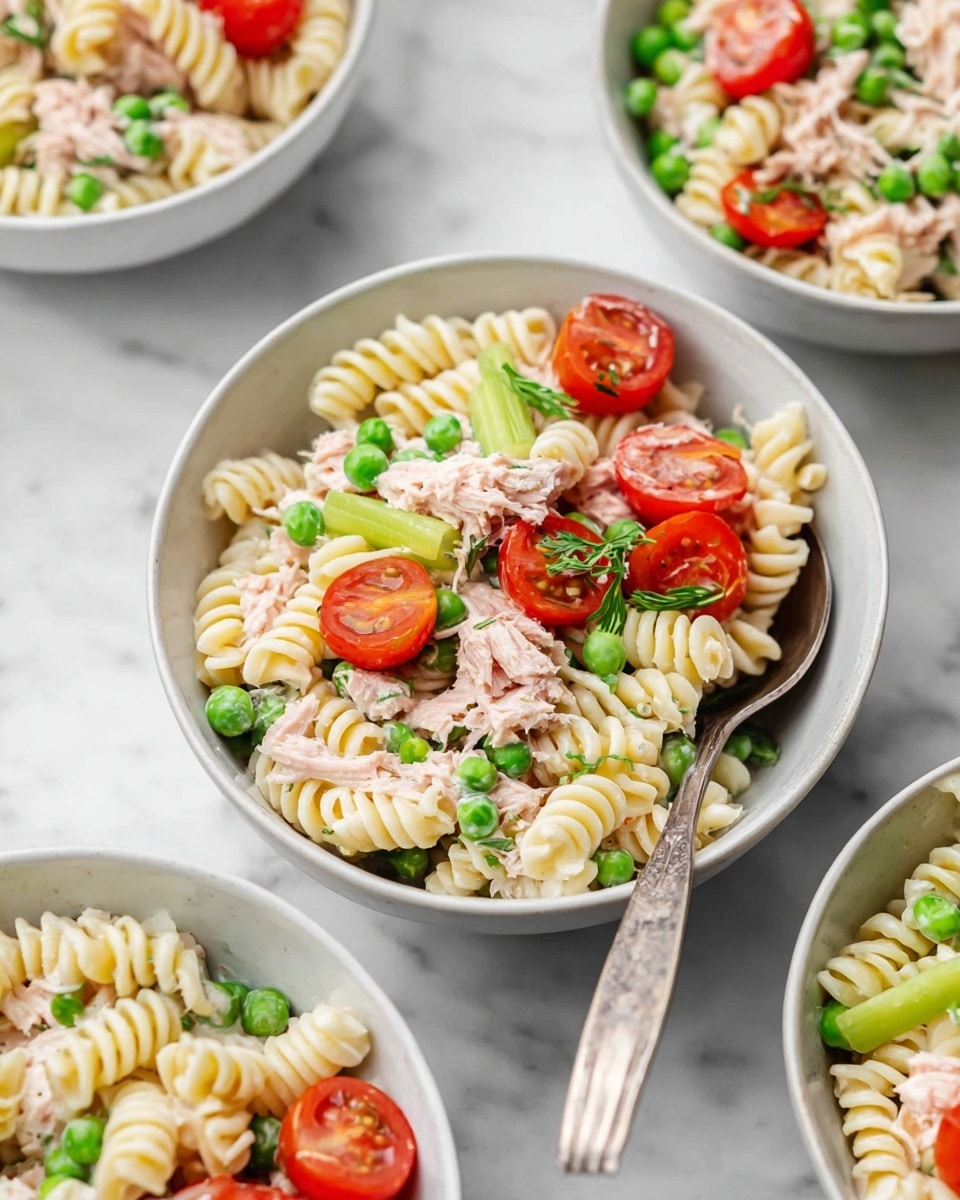 The image shows a white bowl filled with a pasta salad made up of three main layers: twisted pasta spirals that are pale yellow with a soft texture form the base, mixed with bright green peas scattered throughout; pockets of light pink shredded tuna are evenly spread on top, adding a flaky texture; and vibrant red halved cherry tomatoes paired with slices of pale green celery sit on top, adding fresh color and crunch. There is a silver spoon resting inside the bowl, and the bowl is placed on a white marbled surface with other bowls of the same salad partially visible around it. Photo taken with an iphone --ar 4:5 --v 7