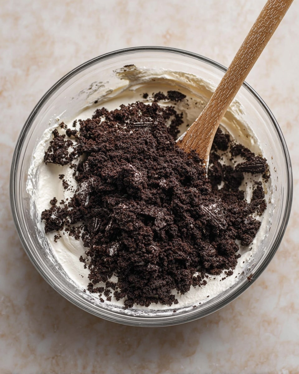 A clear glass bowl sits on a white marbled surface filled with a thick white creamy layer covering the bottom and sides of the bowl. On top of the cream, there is a generous heap of dark brown crushed cookie pieces that give a rough texture with some bigger chunks visible. A wooden spoon is stuck into the mixture, slightly leaning to the side, with its textured light wood grain visible. The overall look is a mix of smooth creamy base and crumbly dark cookie topping in a transparent bowl photo taken with an iphone --ar 4:5 --v 7