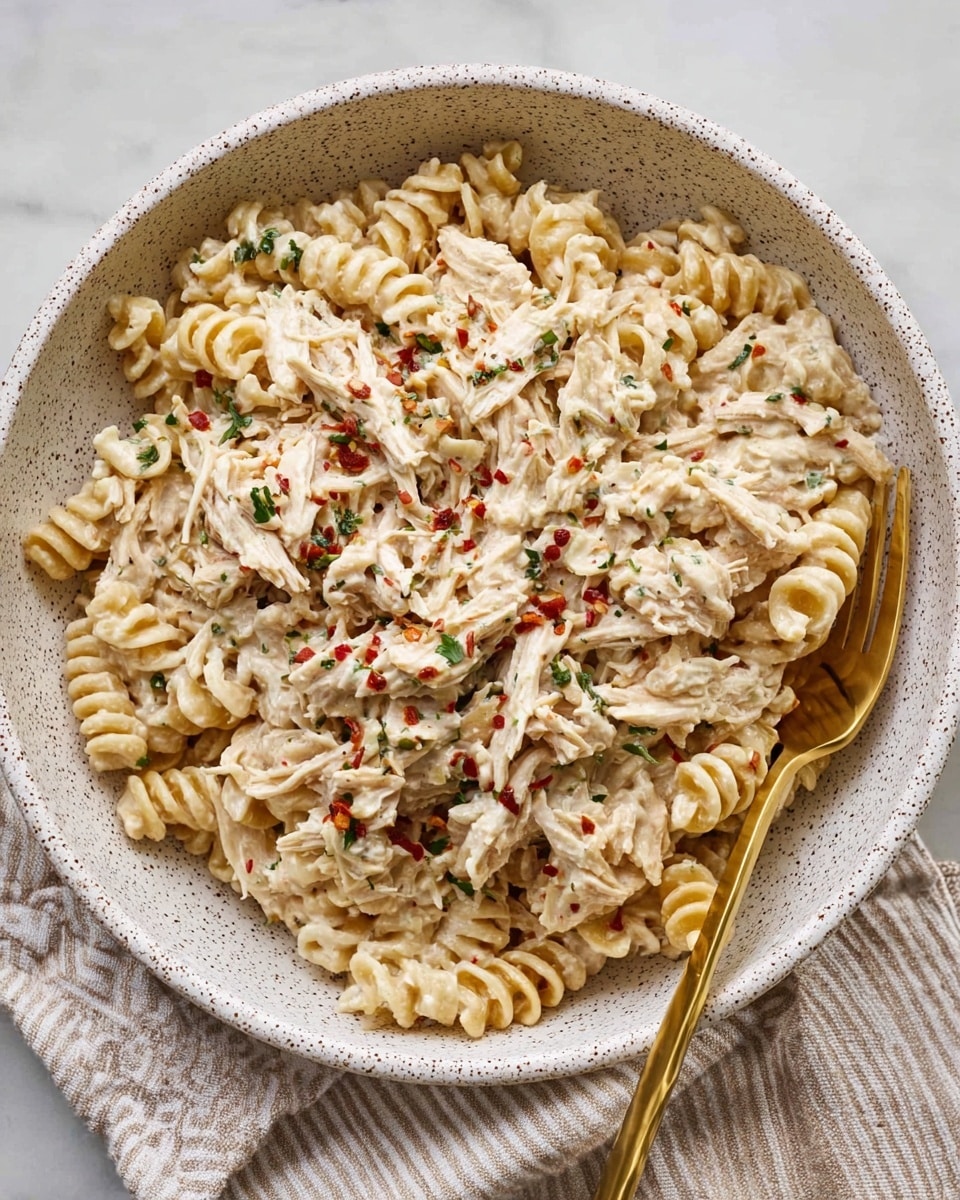 A large white speckled bowl filled with creamy pasta. The dish has about two layers: the bottom layer is short, spiral-shaped pasta with a light beige color, and the top layer is shredded chicken mixed with a white creamy sauce with visible herbs and red chili flakes sprinkled throughout. A silver fork with a gold handle rests inside the bowl on the right side. The bowl is placed on a beige and white striped cloth, all on a white marbled surface. Photo taken with an iphone --ar 4:5 --v 7