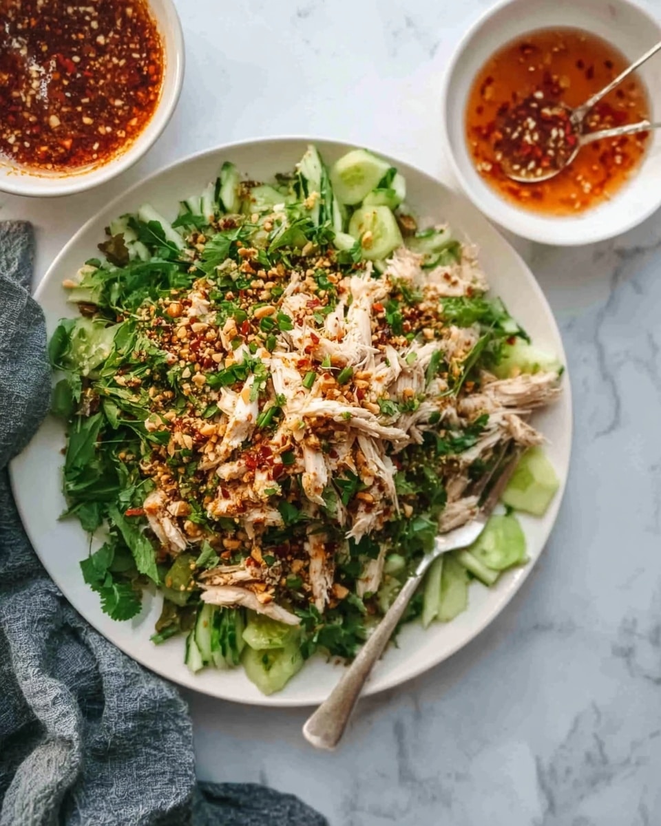 The image shows a white plate filled with a dish that has multiple layers starting with a base of leafy green herbs and thin cucumber strips arranged around the edges. On top of this, there are shredded pieces of cooked chicken with a light brown and white color. The dish is sprinkled with finely chopped green herbs, crushed nuts, and red chili flakes, creating a textured and colorful topping. Near the plate, there is a small white bowl containing a reddish sauce with visible seeds and a spoon inside. The background is a white marbled surface with a woman's hand holding a gray cloth nearby. Photo taken with an iphone --ar 4:5 --v 7