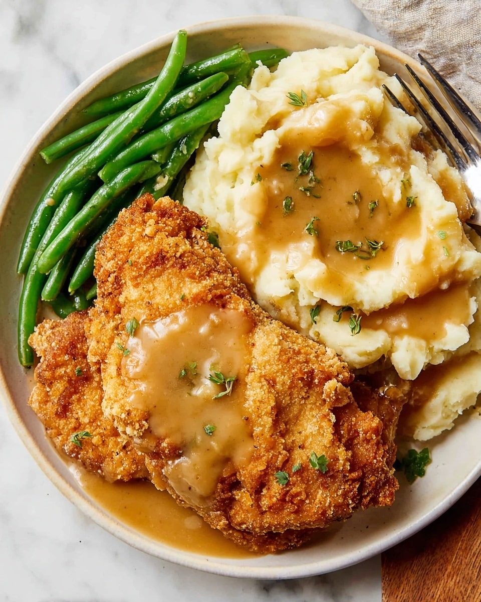 A white plate on a white marbled surface holds a meal with three parts: on the left are bright green beans, in the middle are two large pieces of crispy golden brown fried fried with a rough, crunchy texture, and on the right is a mound of creamy, smooth mashed potatoes topped with thick, light brown gravy poured over both the fried pieces and potatoes. Small green herb bits are sprinkled on top. A fork is placed in the mashed potatoes at the top right side of the plate photo taken with an iphone --ar 4:5 --v 7