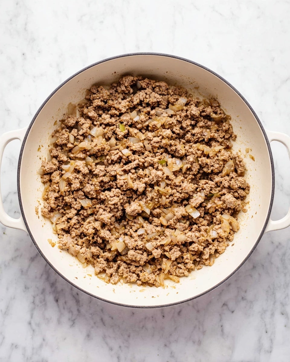 The image shows a white pan filled with browned minced meat mixed with small pieces of translucent cooked onions. The meat is evenly spread inside the pan, with some browned bits visible at the edges. The pan has two handles, one on each side, and it sits on a white marbled surface. The texture of the meat looks crumbly, and the onions add a slight shine and lighter color contrast. Photo taken with an iphone --ar 4:5 --v 7