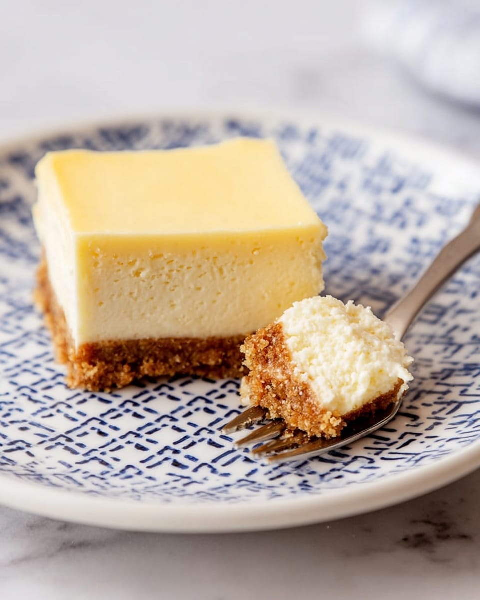 A square piece of cheesecake with two layers rests on a white plate with a blue cross pattern; the top layer is thick, pale yellow, smooth, and creamy while the bottom layer is thin, crumbly, and brown, serving as the crust. To the right, a silver dessert fork holds a small bite-sized piece showing both creamy and crust layers. The plate sits on a white marbled surface. Photo taken with an iphone --ar 4:5 --v 7