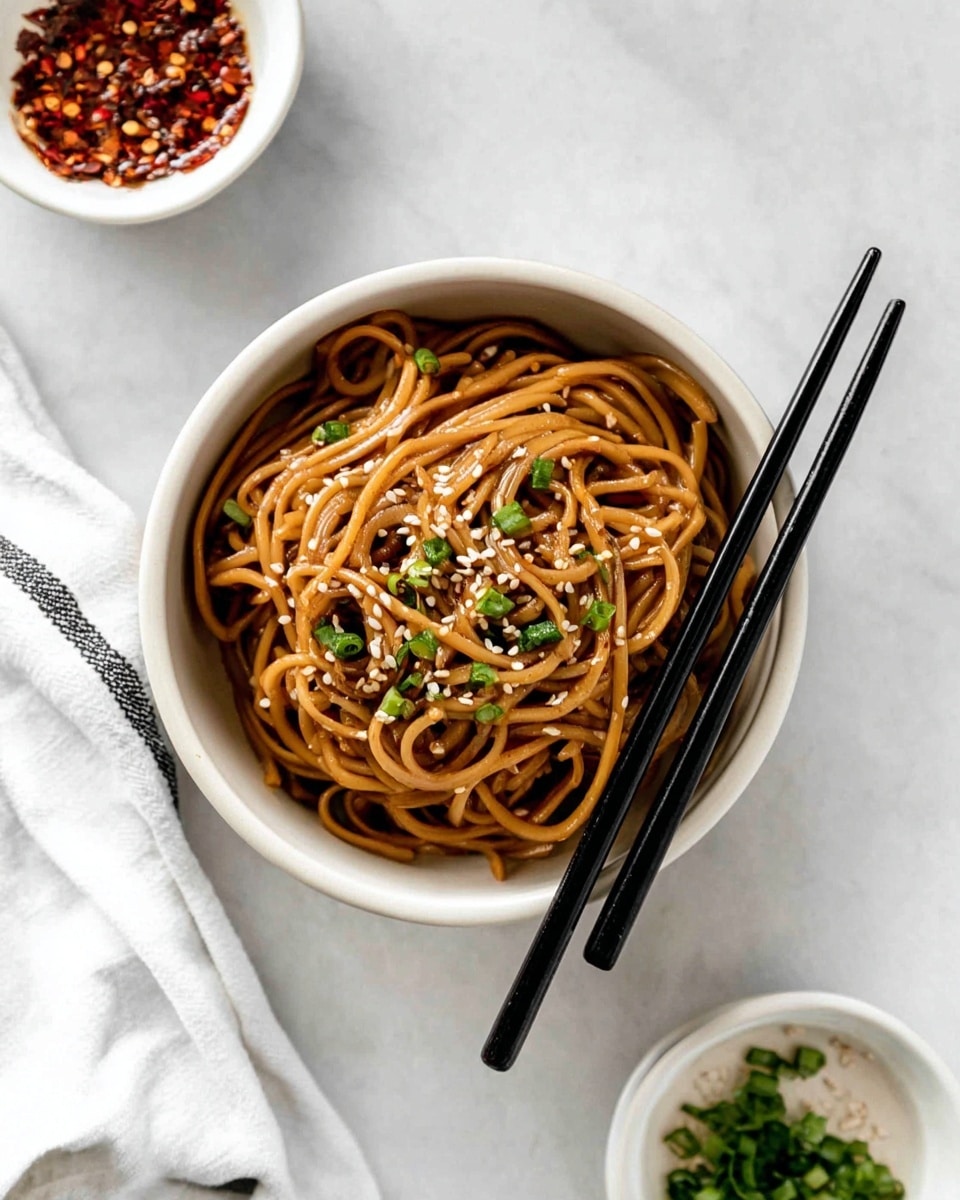 A round white bowl filled with shiny, brown noodles twisted into soft, loose loops. The noodles are topped with small white sesame seeds and bright green chopped chives scattered evenly on top. Two black chopsticks rest diagonally on the bowl’s edge, pointing downwards. In the background on a white marbled surface, two small white bowls hold red chili flakes and more chopped green chives. A white cloth with black stripes lies partially visible next to the bowl. The overall scene is clean and bright. photo taken with an iphone --ar 4:5 --v 7
