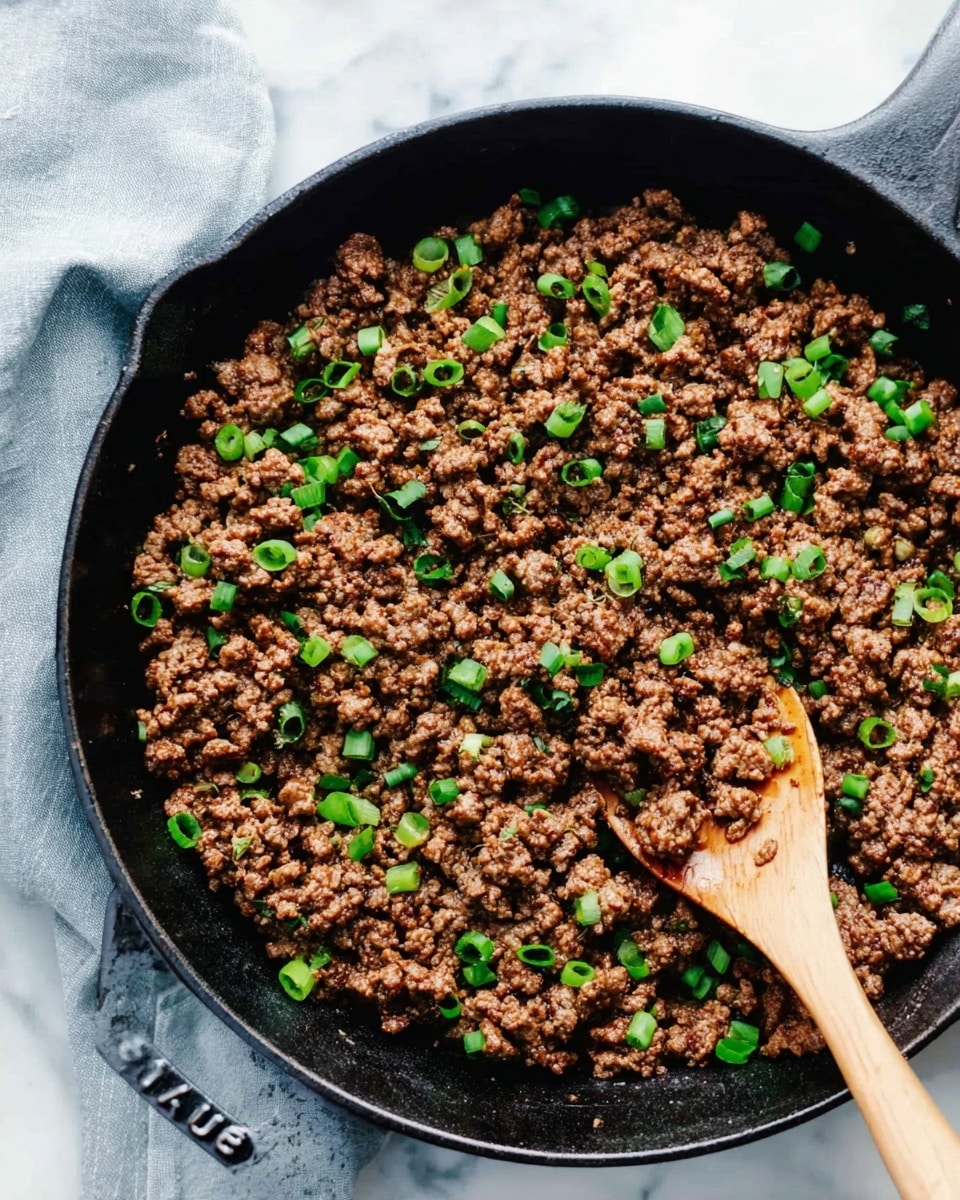 A black cast iron pan filled with cooked ground meat that is brown and crumbly in texture, mixed with evenly scattered sliced green onions which add bright green color on top. There is a wooden spoon resting in the pan with some ground meat on it. The pan is placed on a white marbled surface with a light gray cloth napkin visible in the top left corner. The edges of the pan show the word